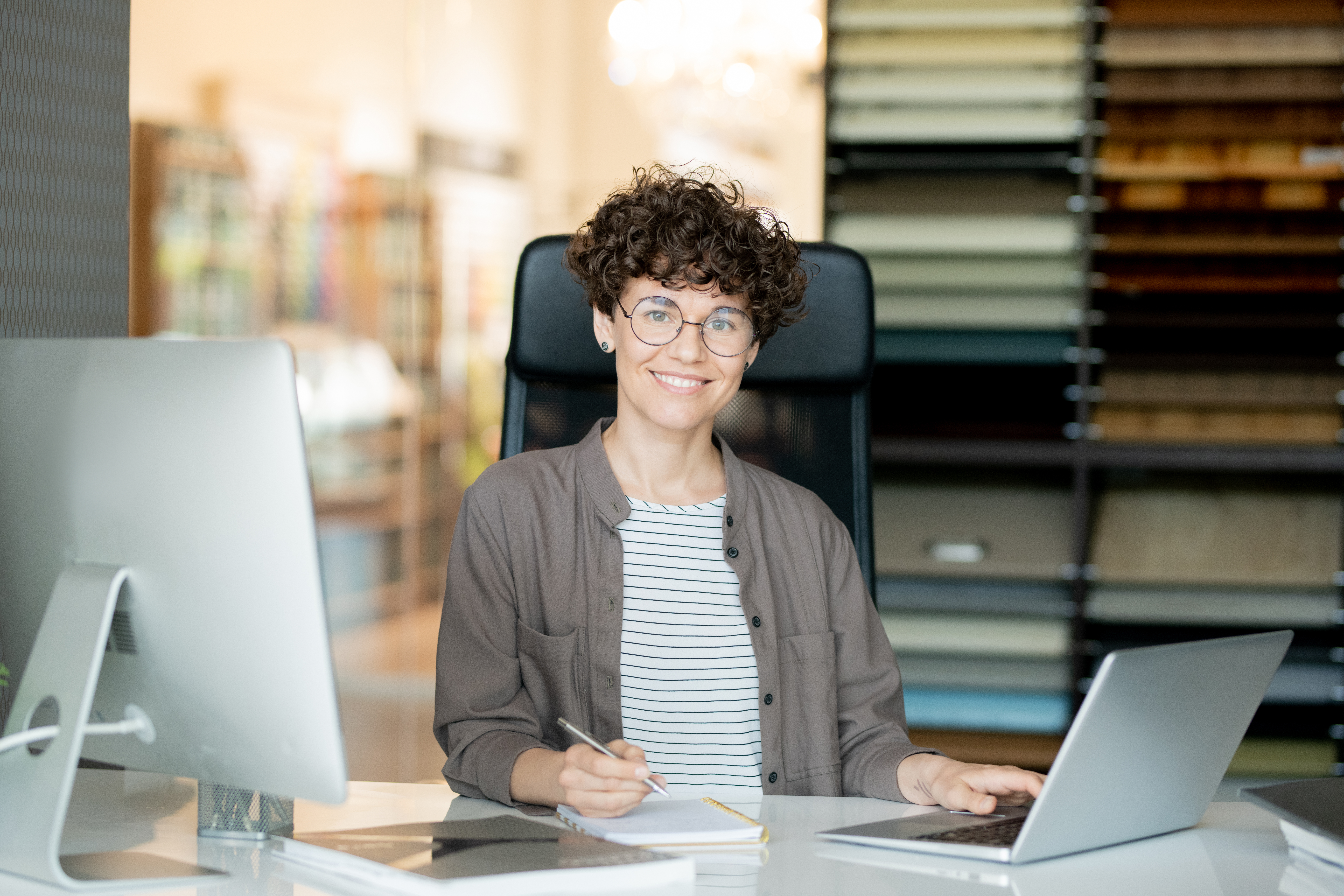 Eine Person mit lockigem Haar und Brille sitzt an einem Schreibtisch, arbeitet am Laptop und lächelt. Im Hintergrund sind Farbproben sichtbar.