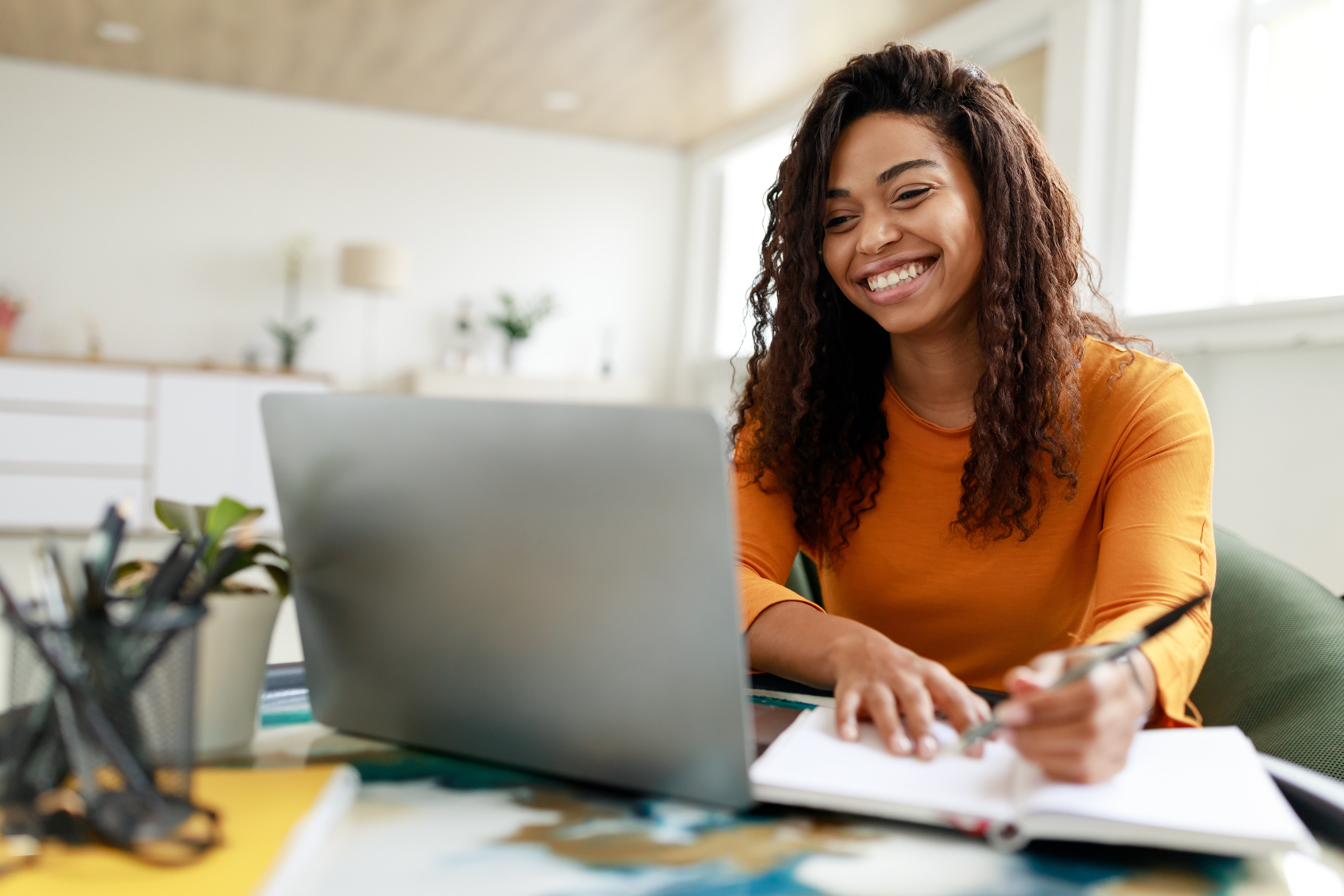 A woman with curly hair sits at a desk, smiling as she works on a laptop. She holds a pen in her hand and writes something in a notebook. Plants and stationery are visible around her.