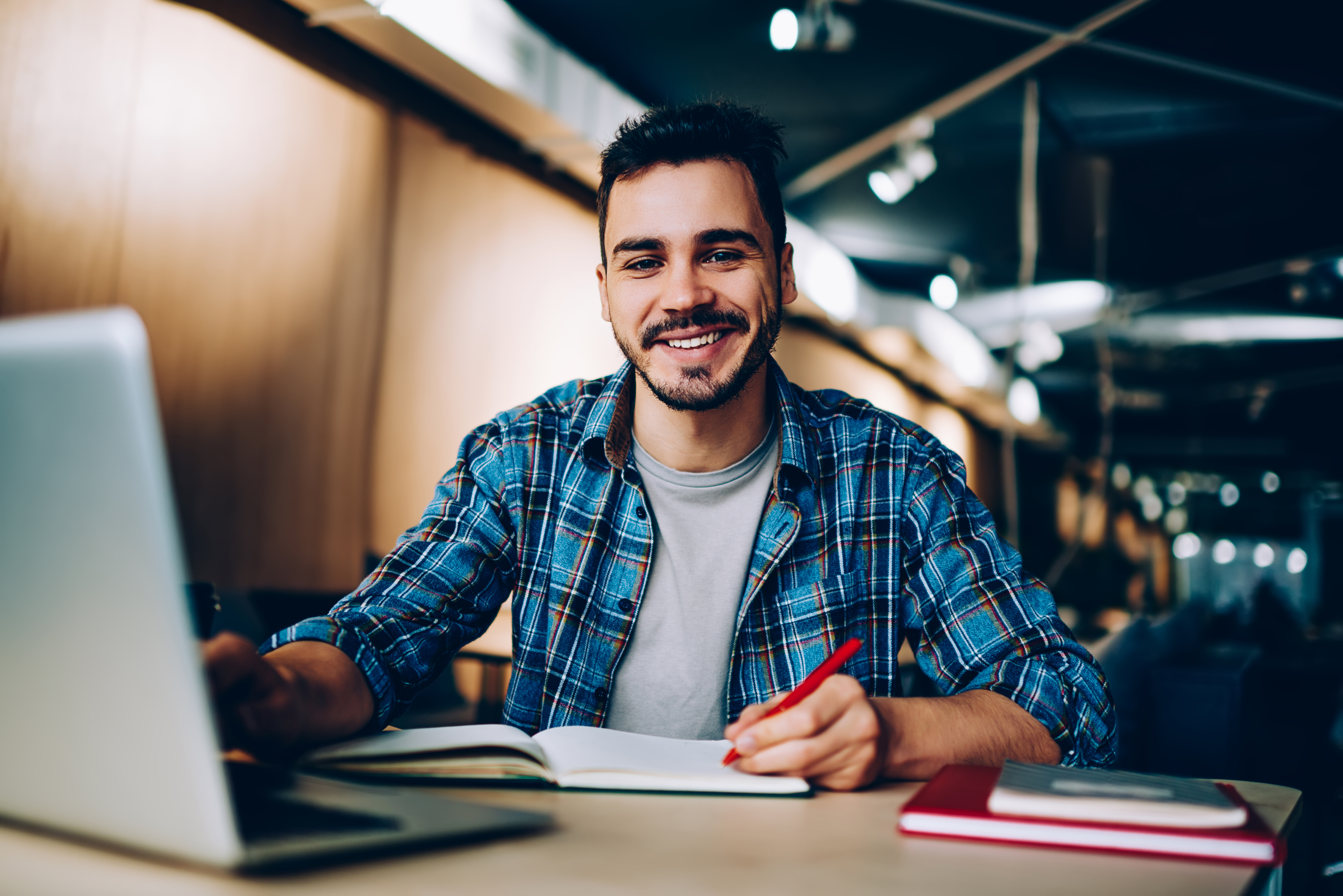 A young man with a beard is sitting at a table, smiling and looking at the camera. He is wearing a checked shirt and holding a red pen. In front of him, there is a notebook and a laptop.