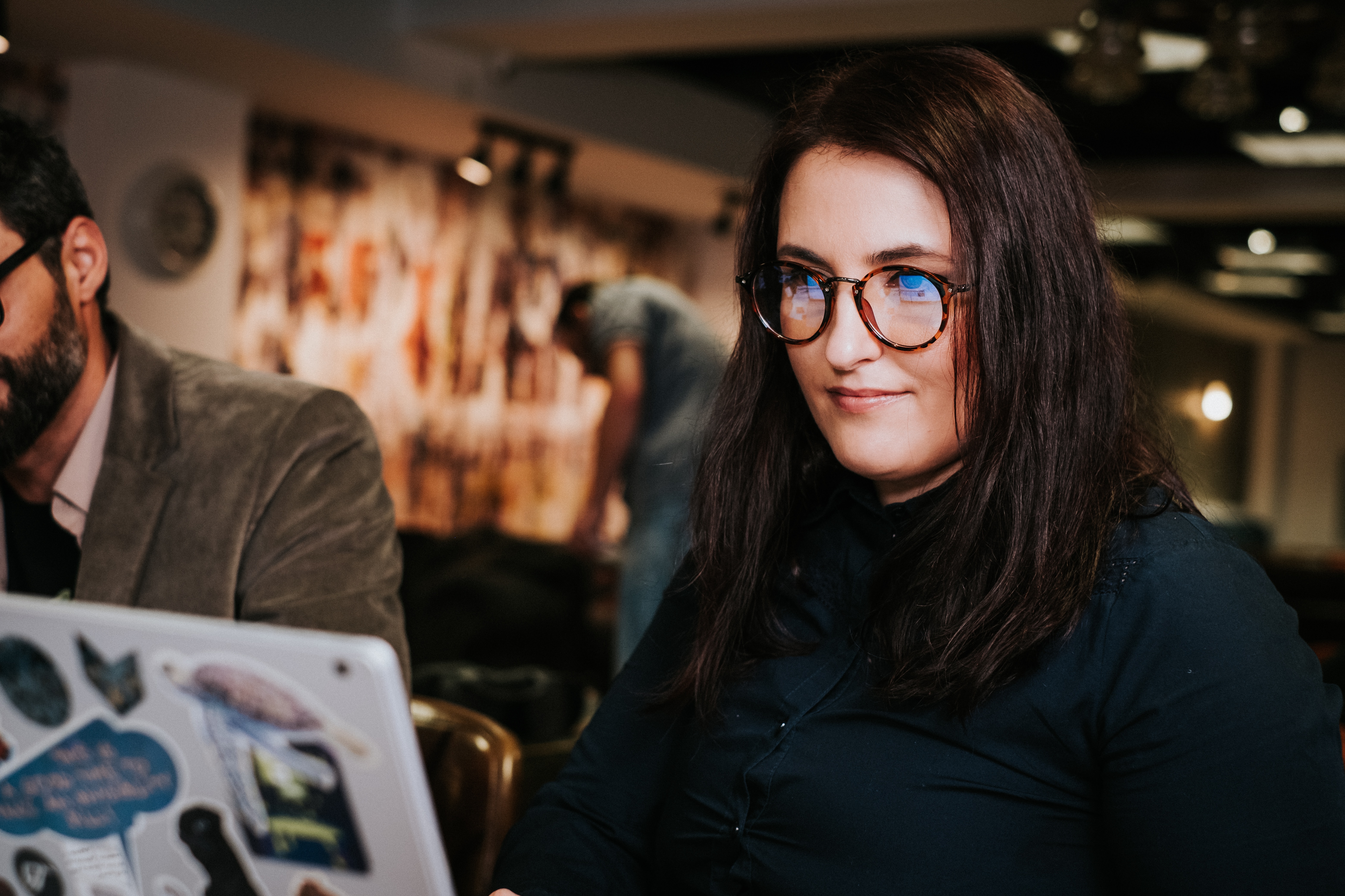 A woman with long, dark hair and glasses is sitting at a table, working on a laptop covered in stickers. In the background, a man can be seen sitting at another table.