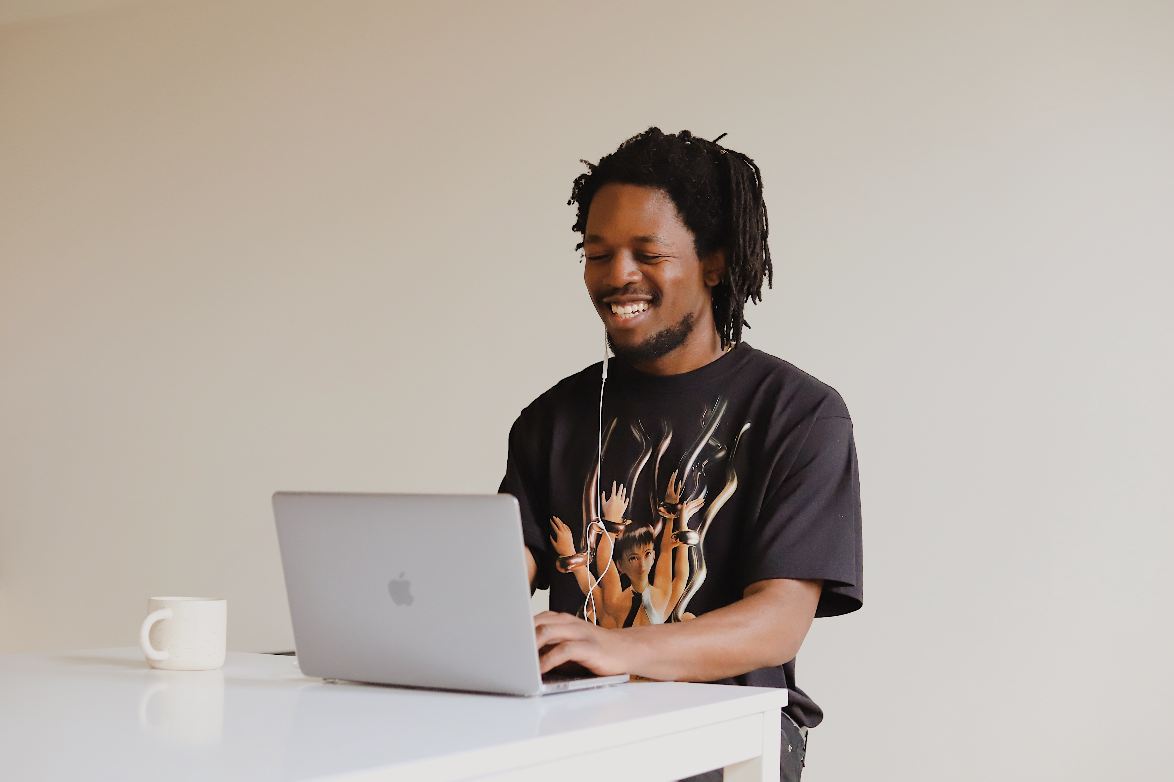 A man with curly hair is sitting at a table, working on a laptop. He is wearing a T-shirt with an artistic design. Next to him, there is a cup. The room has a light, neutral wall color.