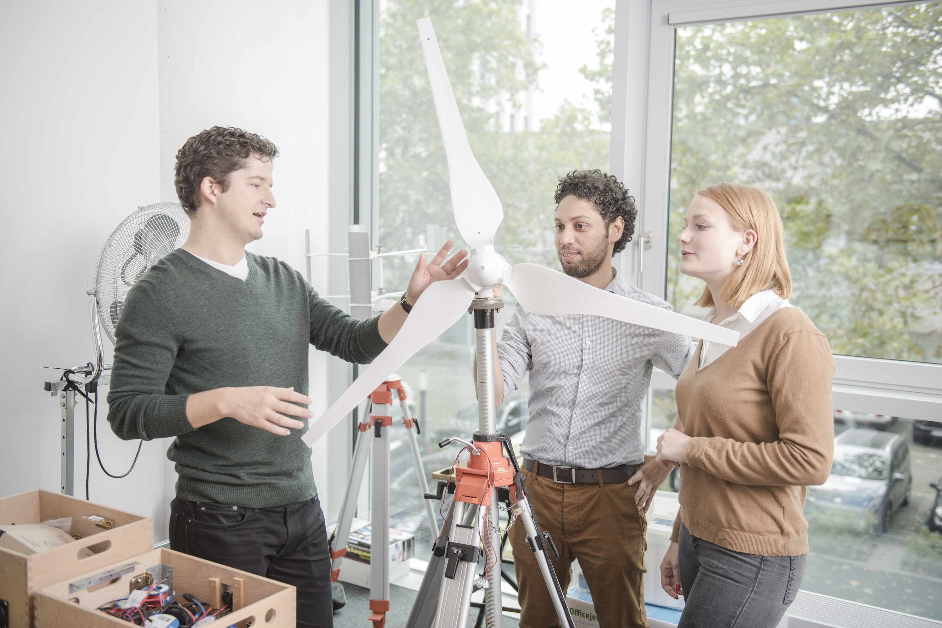 Three people are standing in a room, looking at a model of a wind turbine. One person is explaining how it works while the others listen attentively. Tools and materials are visible in the background.