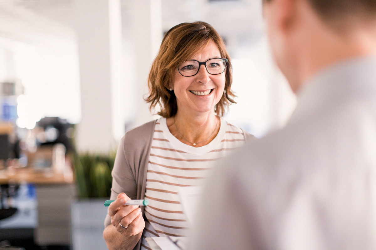 Eine lächelnde Frau mit Brille trägt ein gestreiftes T-Shirt und einen grauen Cardigan. Sie hält einen Stift in der Hand und spricht mit einer anderen Person in einem modernen Büro. Pflanzen sind im Hintergrund sichtbar.
