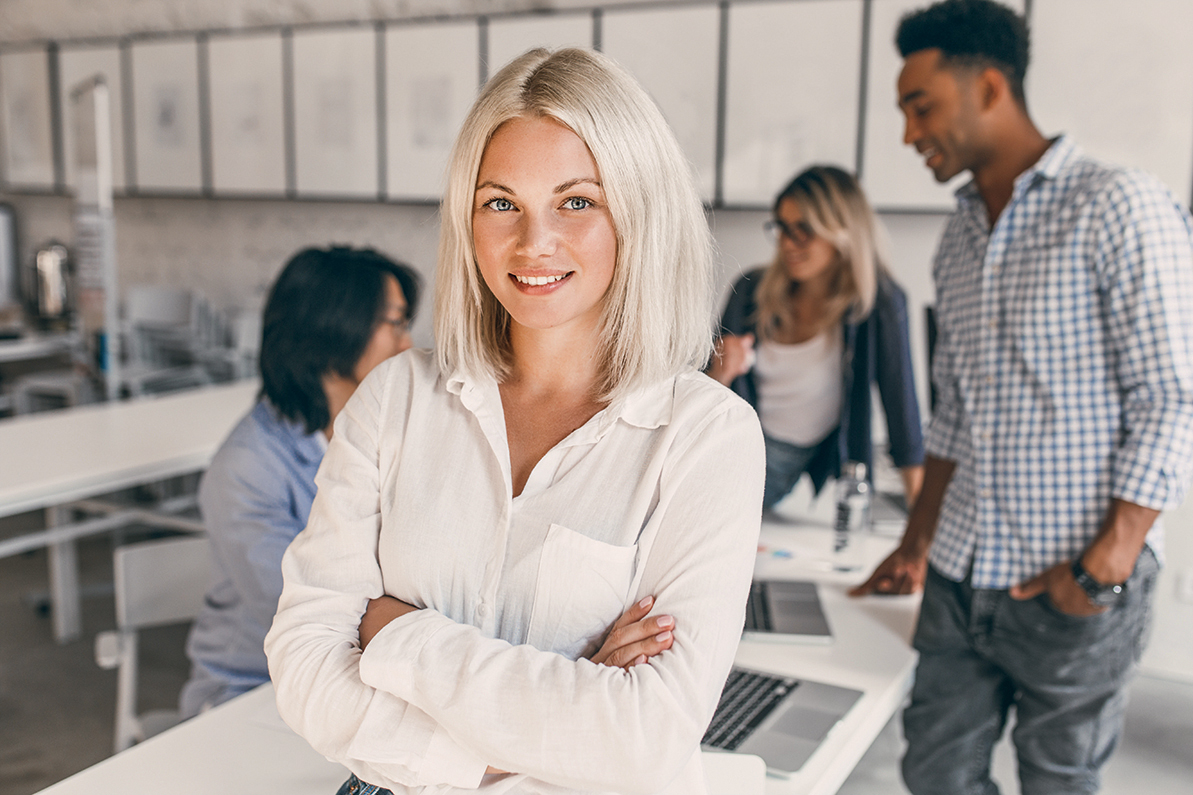 Eine blonde Frau mit kurzen Haaren steht lächelnd im Vordergrund, während drei Personen im Hintergrund an einem Tisch arbeiten. Alle sind in einem modernen Büro mit Laptops und Notizen beschäftigt.