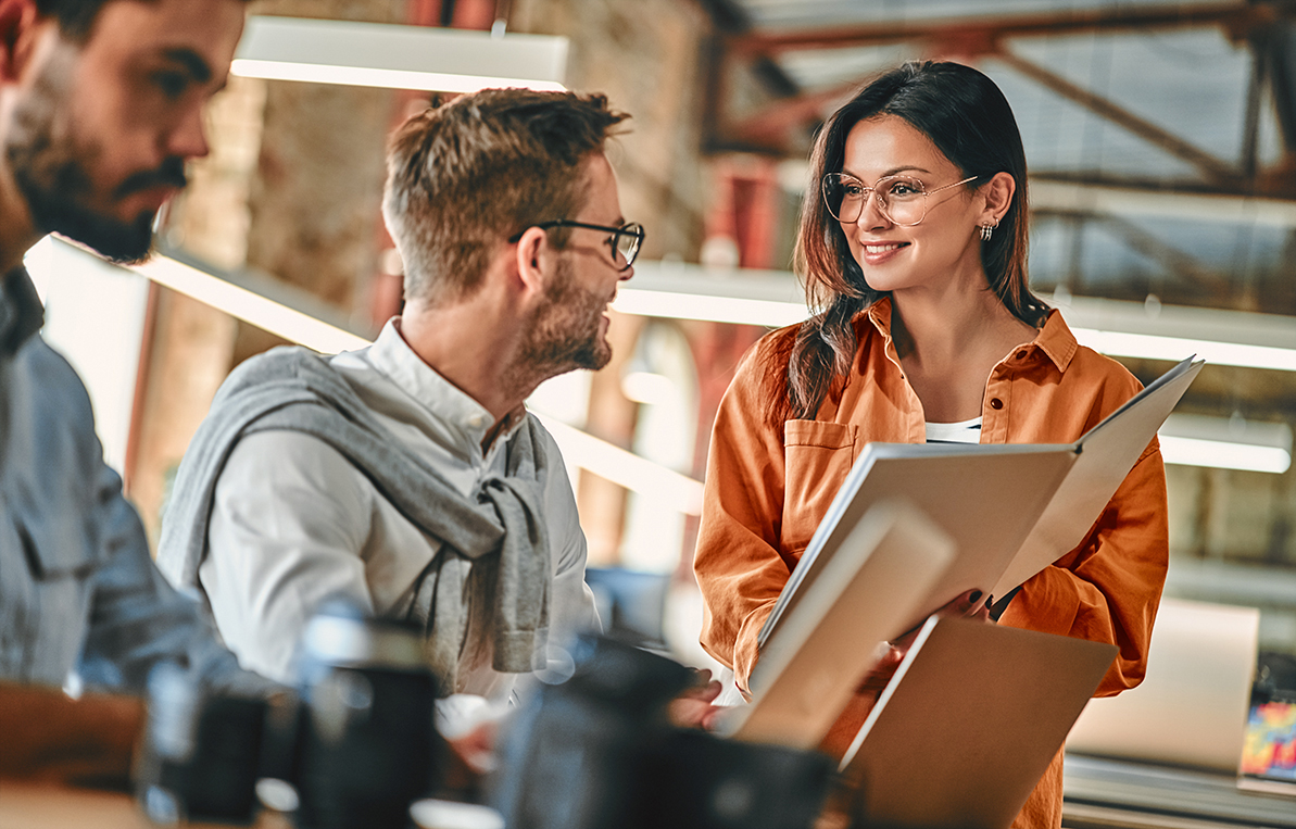 Eine Frau mit Brille hält ein Dokument und spricht mit einem Mann, während ein weiterer Mann am Tisch sitzt. Alle drei scheinen an einem kreativen Projekt zu arbeiten. Der Raum hat eine moderne, industrielle Atmosphäre.