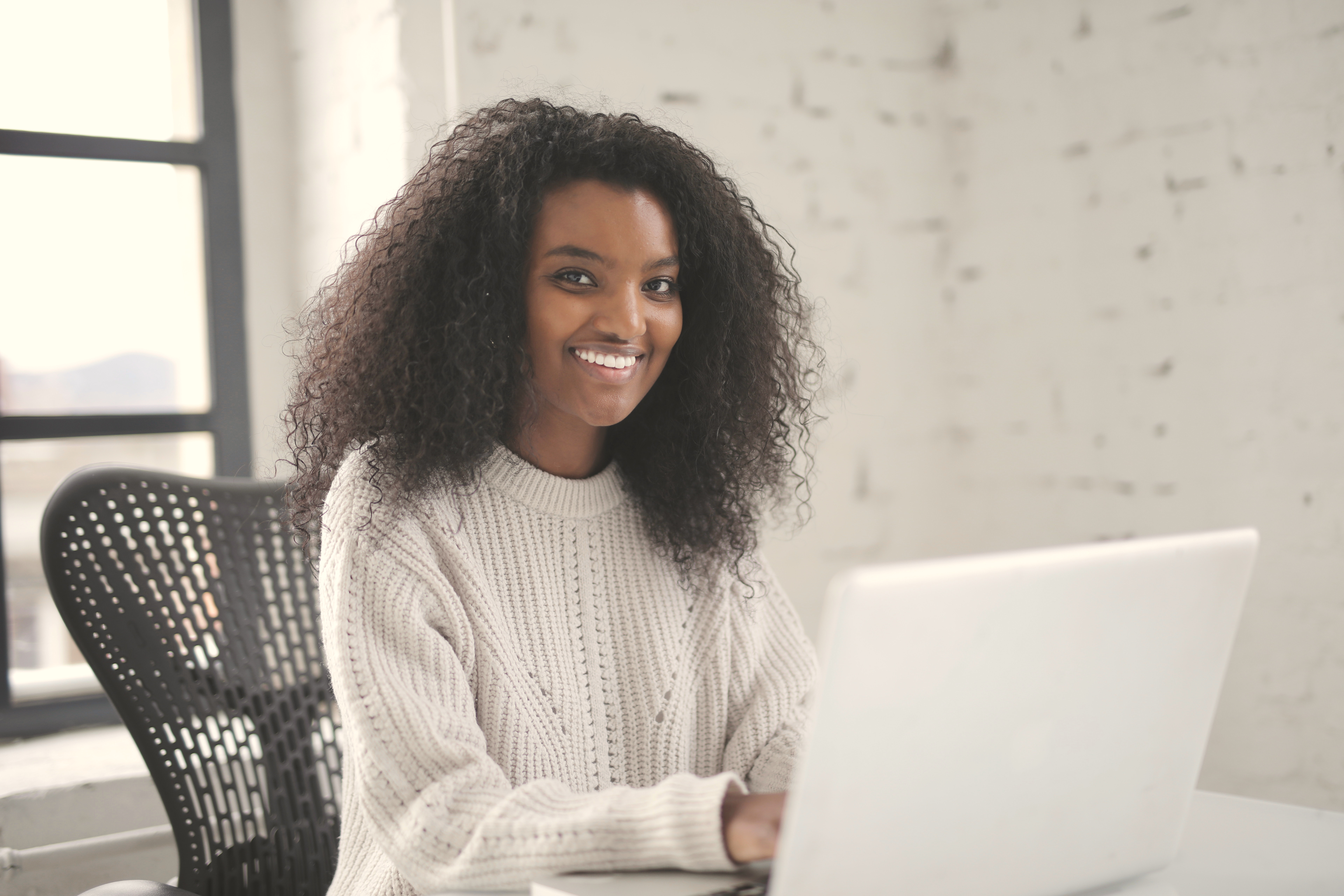 A person with curly black hair is sitting at a desk, working on a laptop. They are wearing a light, knitted sweater and smiling at the camera. The background features a bright, minimalist setting.