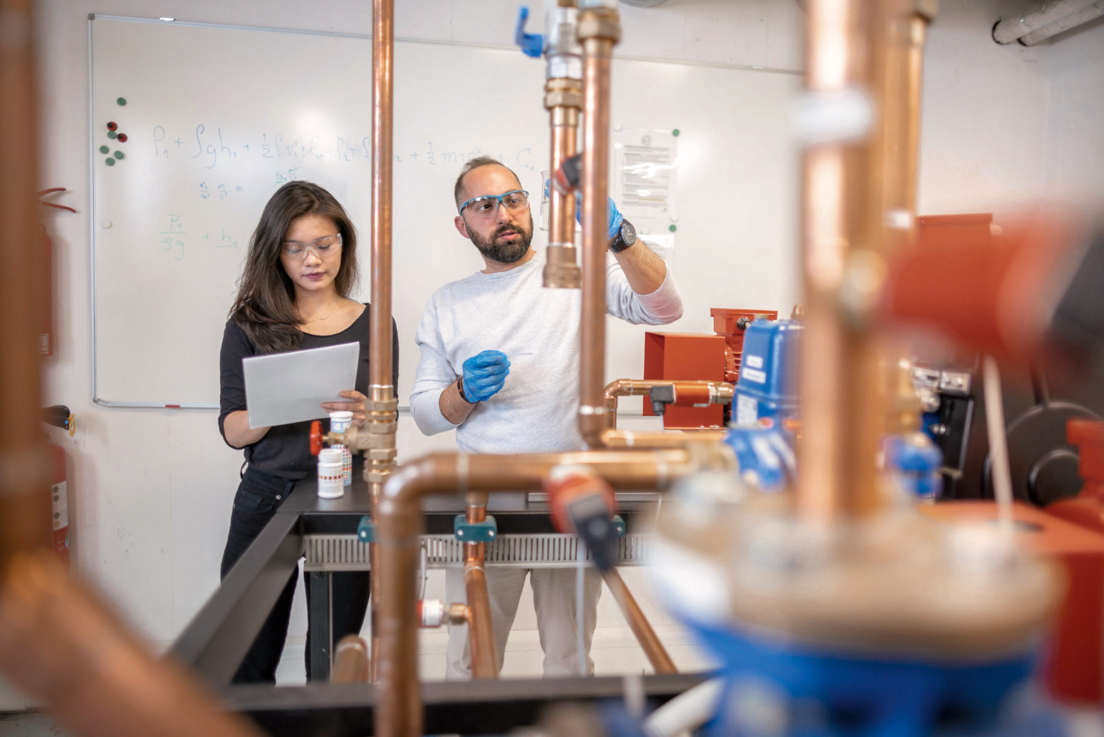 A man and a woman are standing in a laboratory, working on a copper pipe installation. The man is holding a pipe and explaining something, while the woman is taking notes on a sheet of paper. They are wearing safety gear.