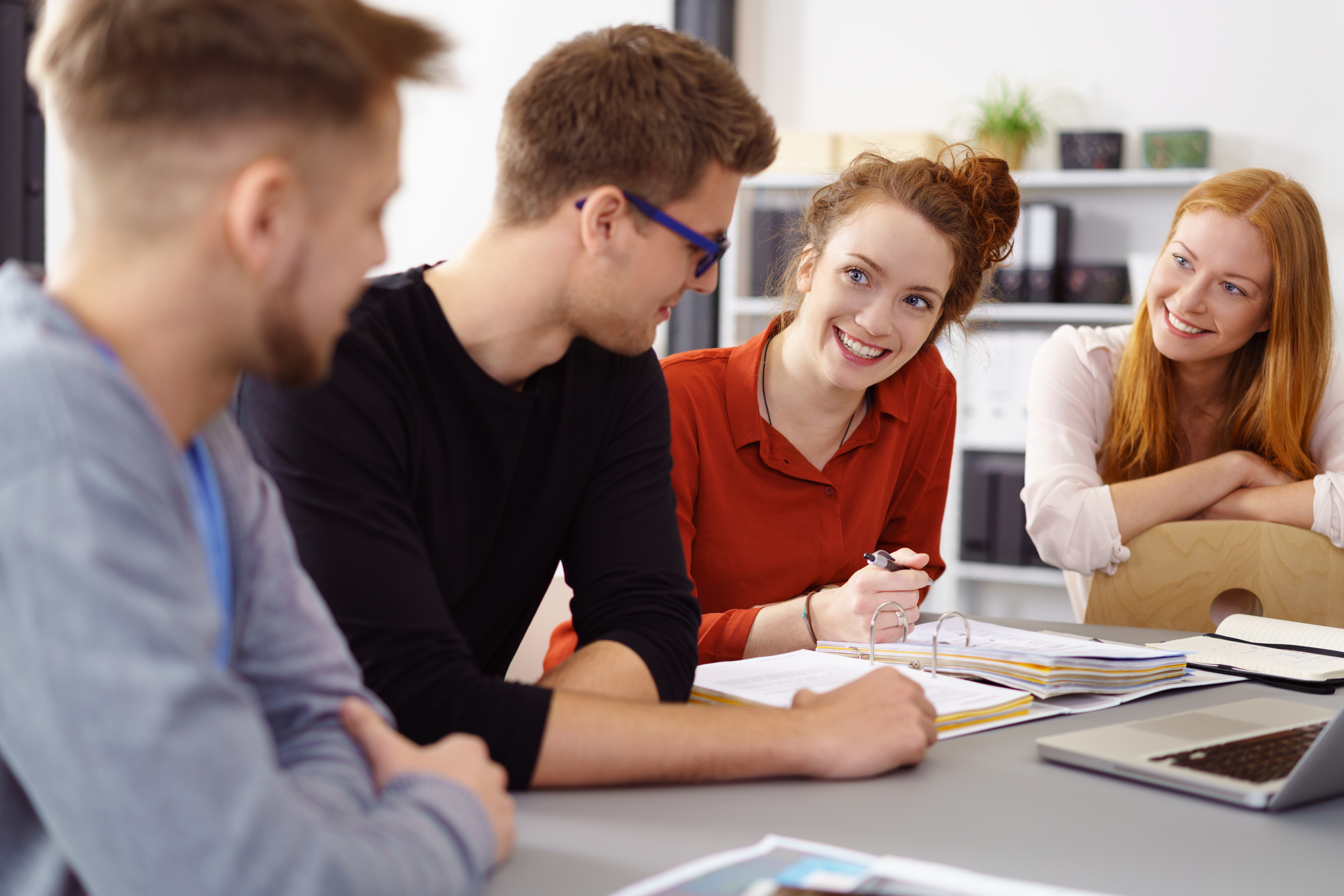 Four young adults are sitting at a table, discussing and smiling. Two men and two women are present, with notes and a laptop in front of them. The atmosphere feels relaxed and collaborative.