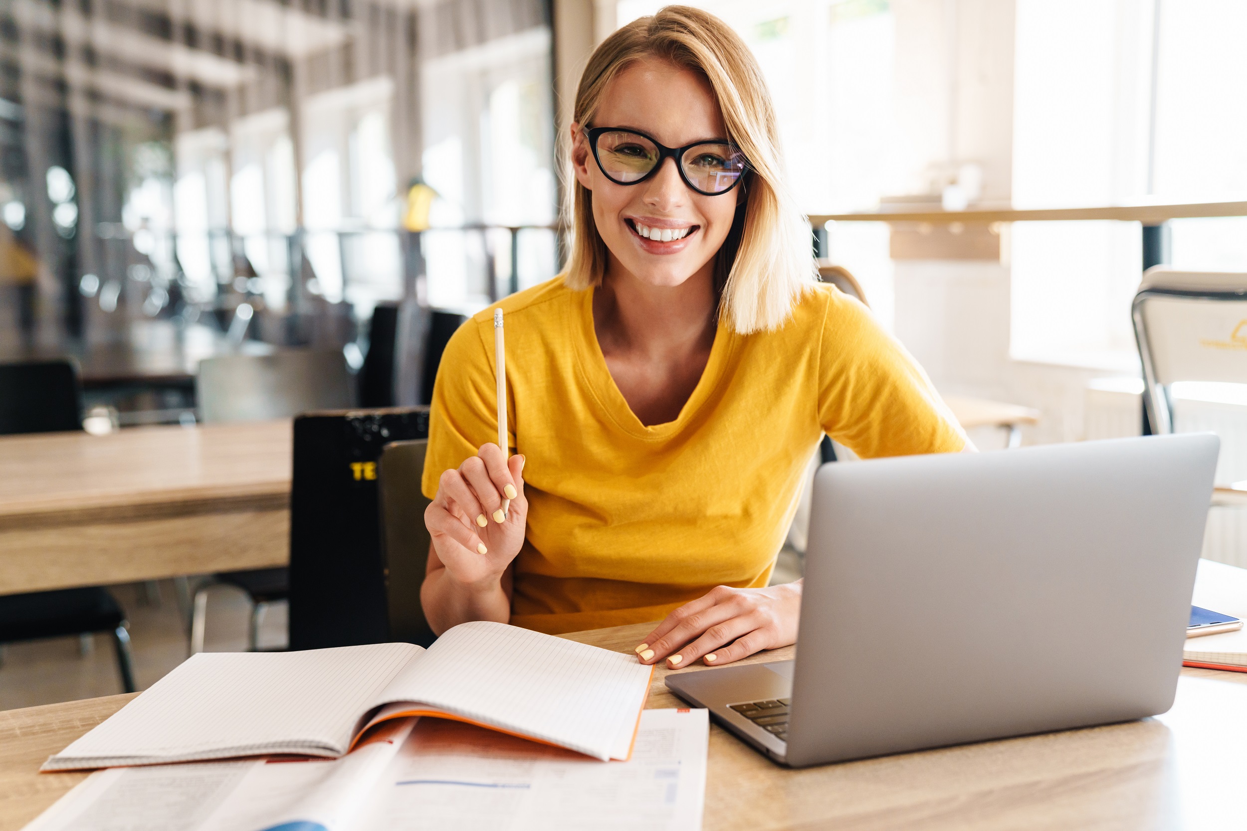 Eine lächelnde Frau mit Brille sitzt an einem Tisch, arbeitet an einem Laptop und hält einen Stift in der Hand. Vor ihr liegen aufgeschlagene Bücher und Notizen. Sie trägt ein gelbes T-Shirt.