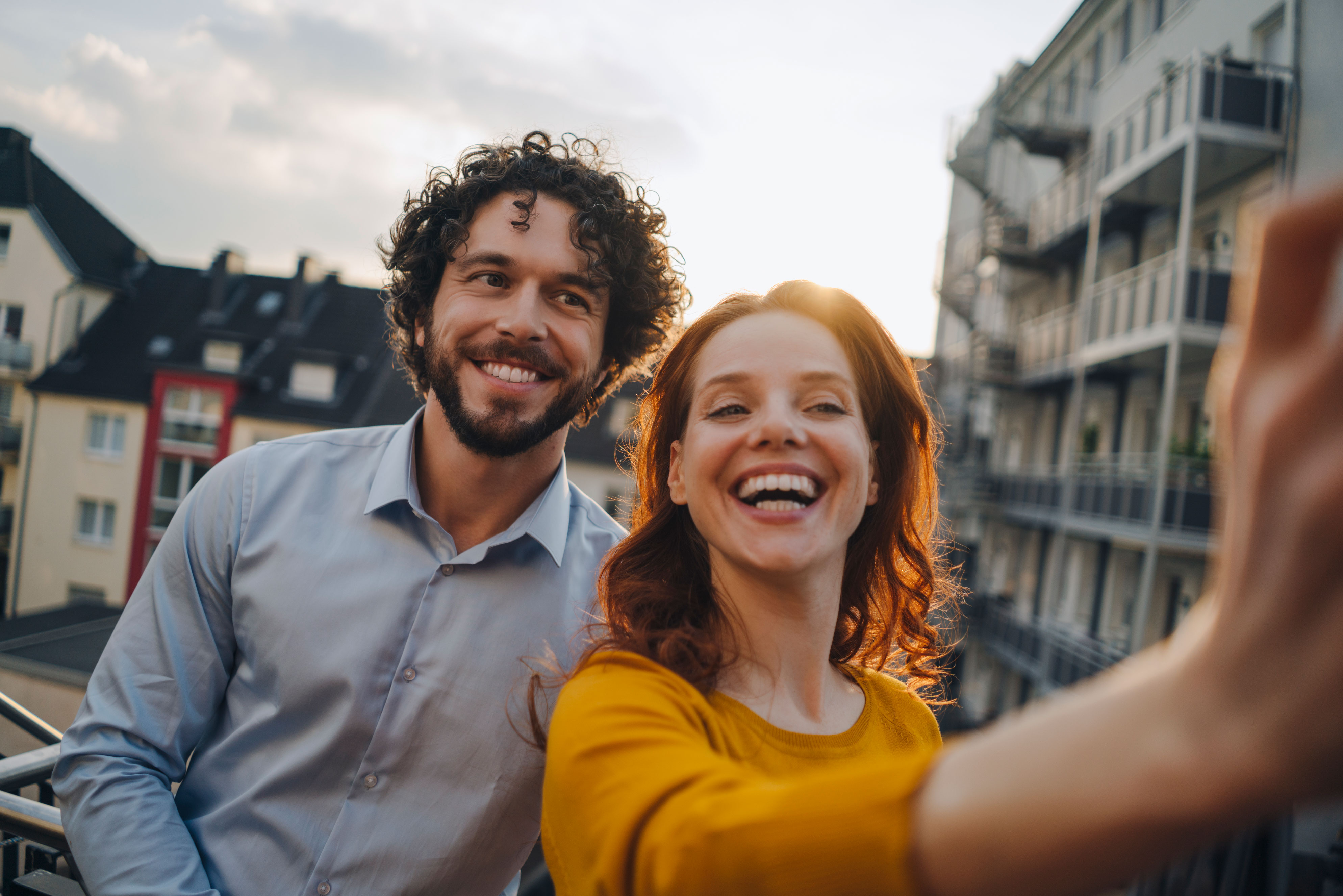 A man with curly hair and a woman with long red hair are smiling as they take a selfie on a balcony. In the background, you can see residential buildings and a cloudy sky.