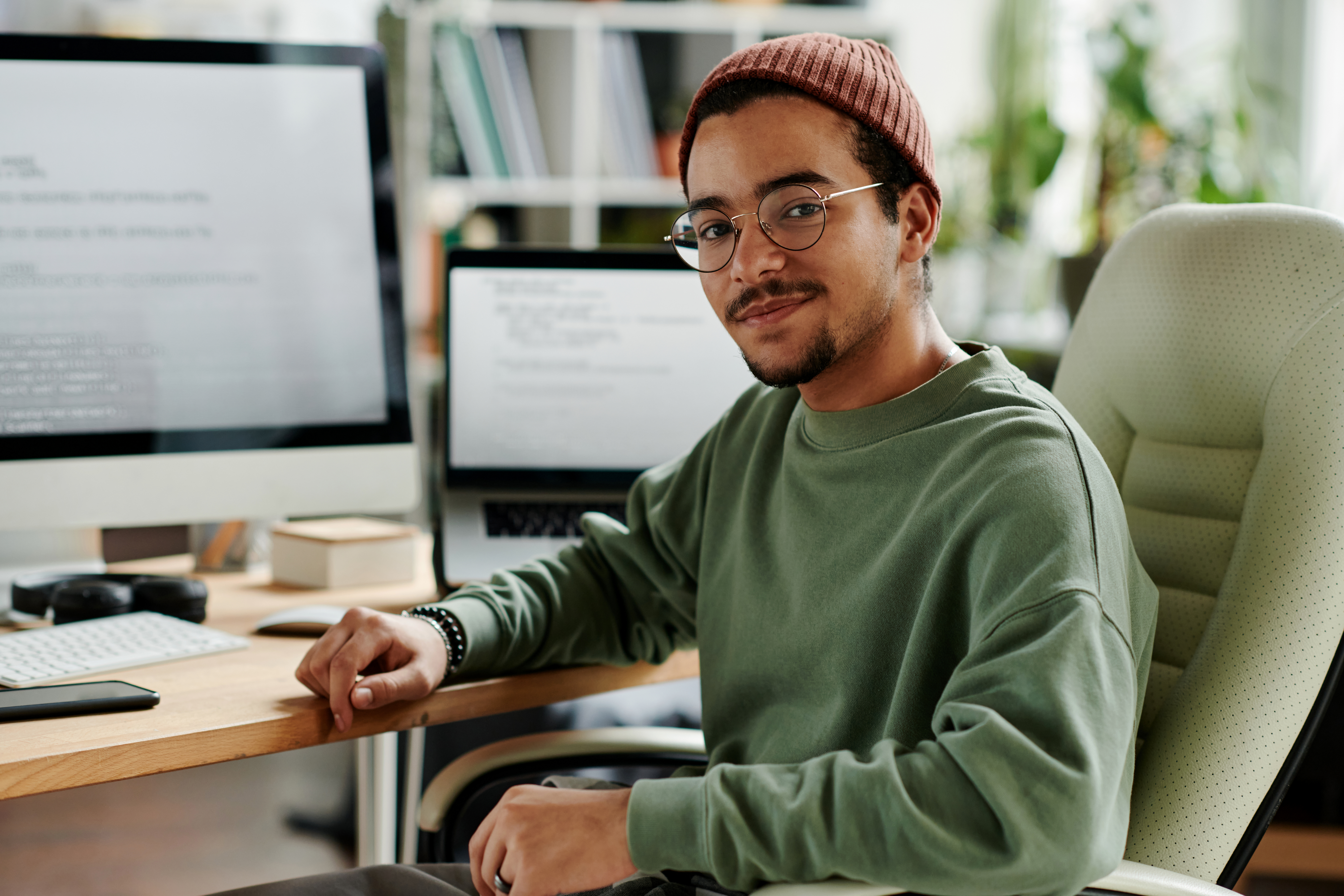 A young man with glasses and a red hat is sitting at a desk. He is wearing a green sweatshirt and smiling. In the background, computer screens and office supplies are visible.