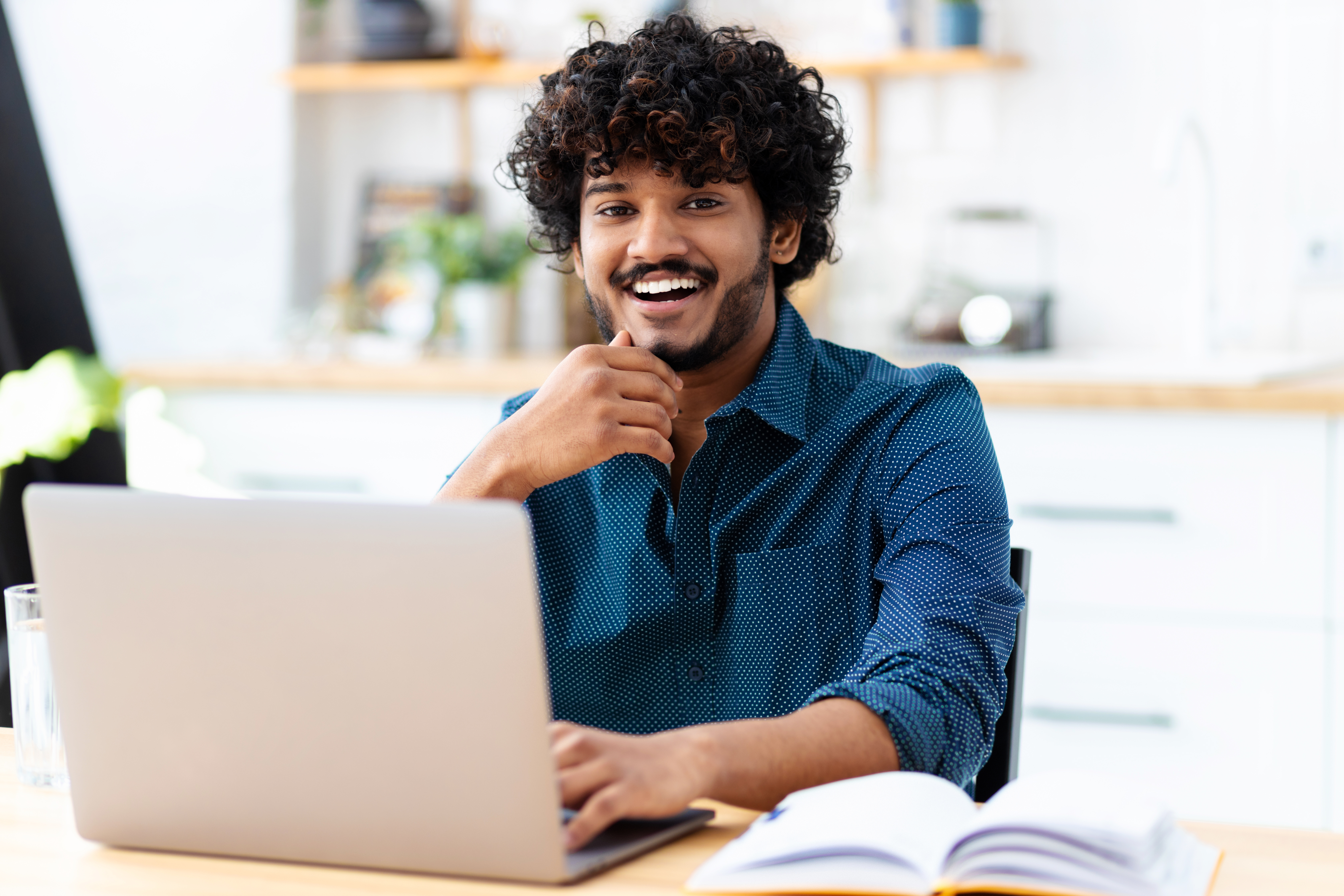 A young man with curly hair sits at a table, smiling as he looks at a laptop. In front of him lies an open book, and a glass of water is placed beside it.