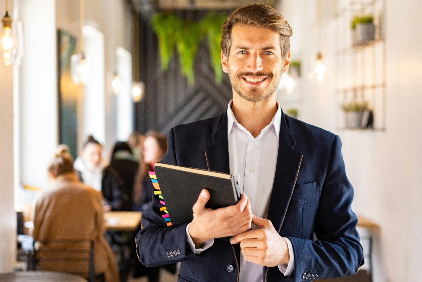 A man in a blue blazer is holding a notebook and smiling. In the background, there are blurred figures in a café. The atmosphere feels friendly and inviting.