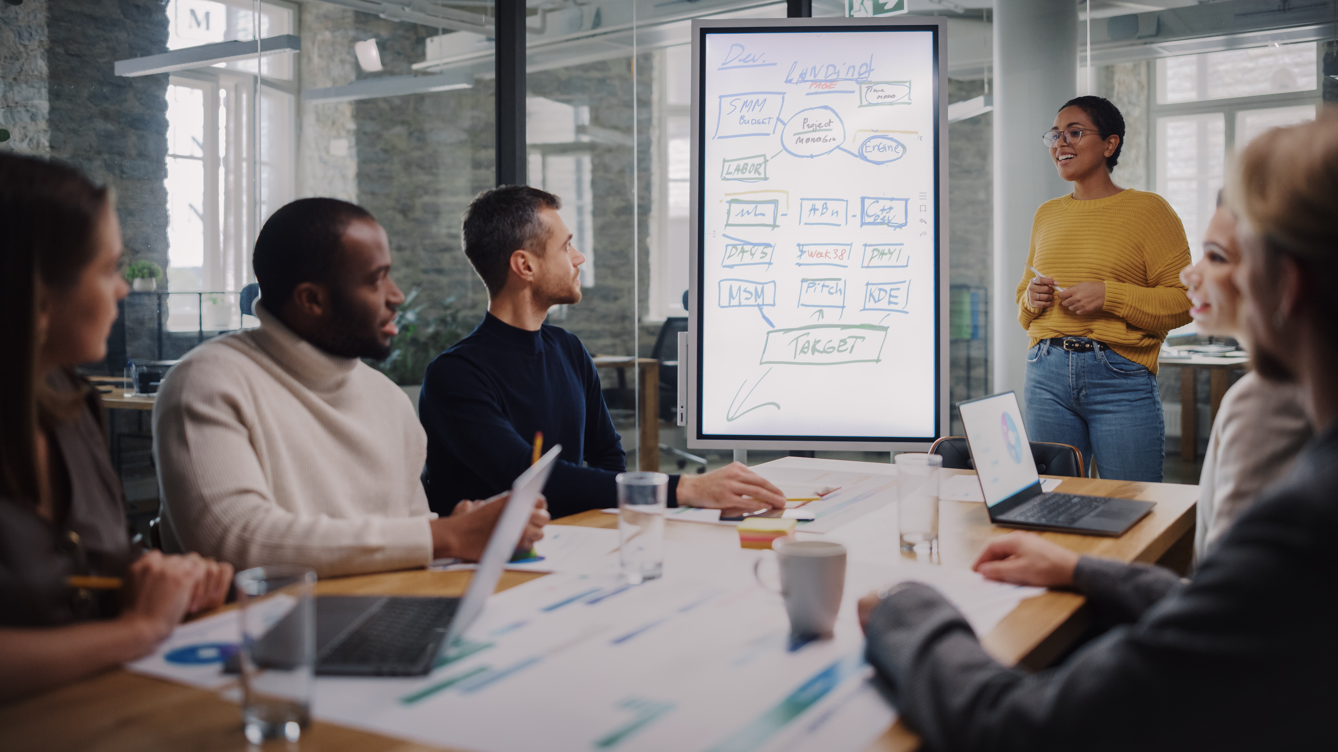 Eine Person steht vor einem Flipchart und präsentiert Informationen. Sechs Personen sitzen an einem Tisch und hören aufmerksam zu. Laptops und Getränke sind auf dem Tisch verteilt, während Notizen und Diagramme sichtbar sind.
