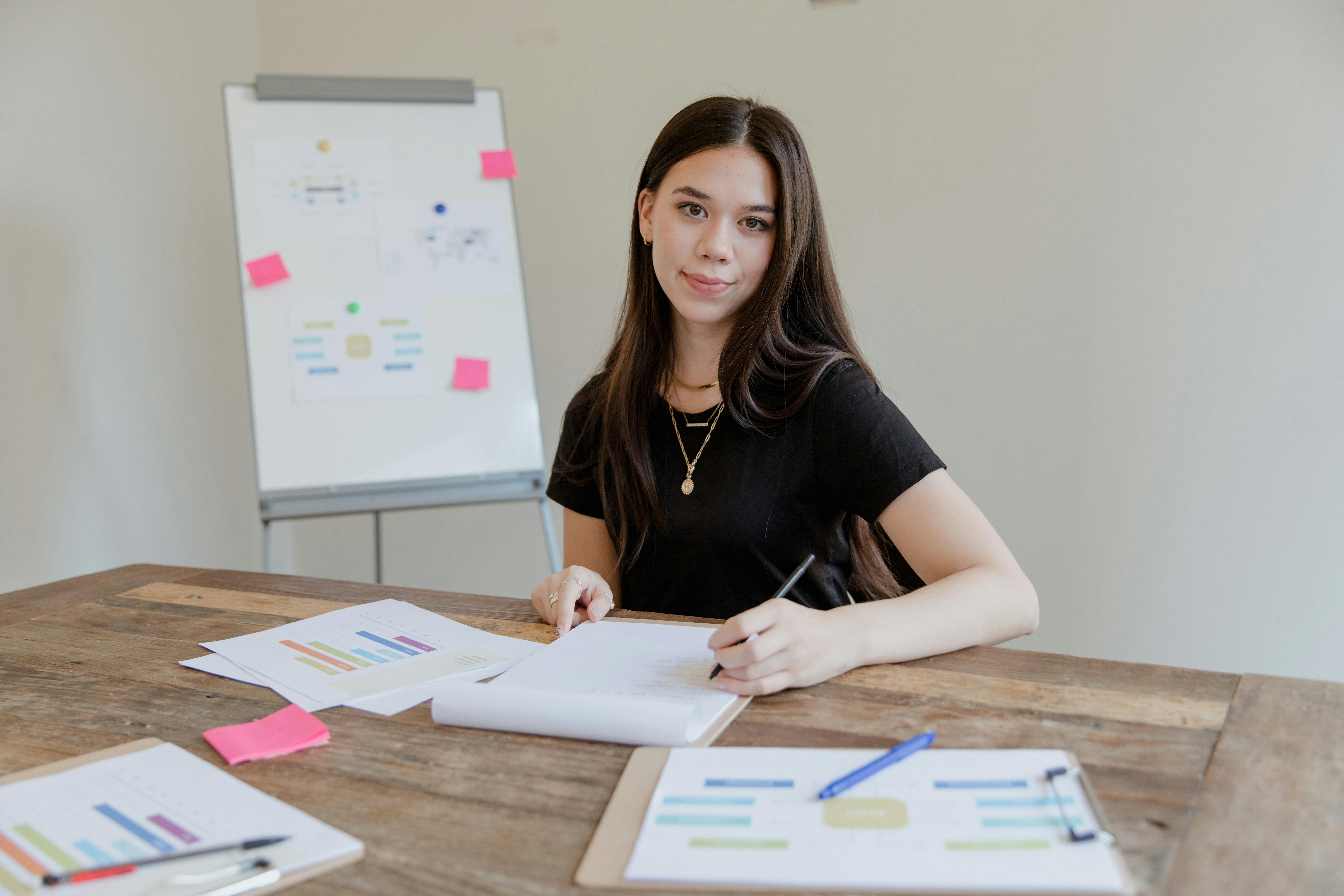 A young woman is sitting at a table, writing in a notebook and looking at the camera. On the table, there are several sheets with diagrams and notes. In the background, there is a flip chart with colorful sticky notes.
