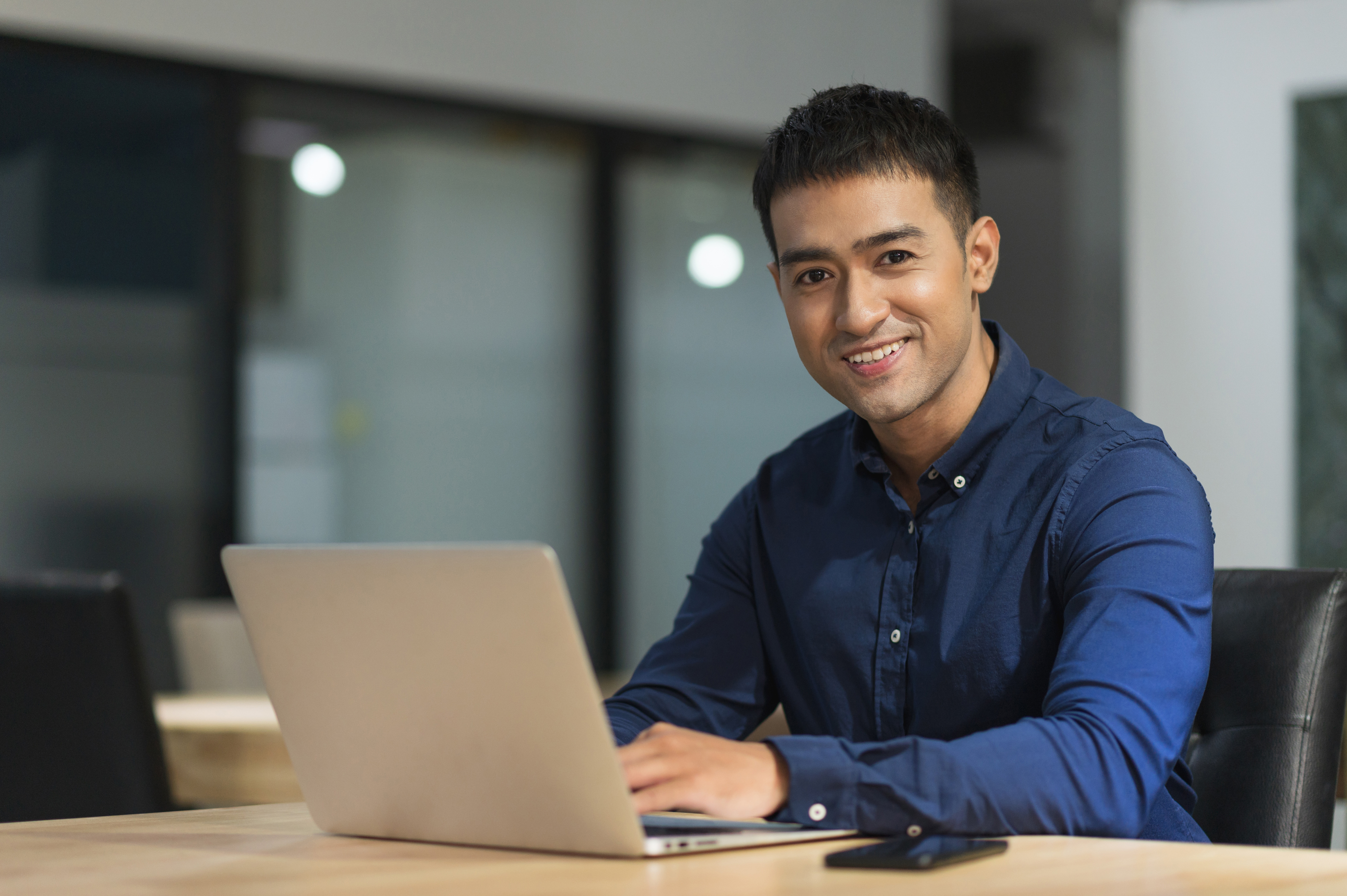 A young man is sitting at a table, working on a laptop. He is wearing a blue shirt and smiling. In the background, there are blurred windows and office furniture visible.
