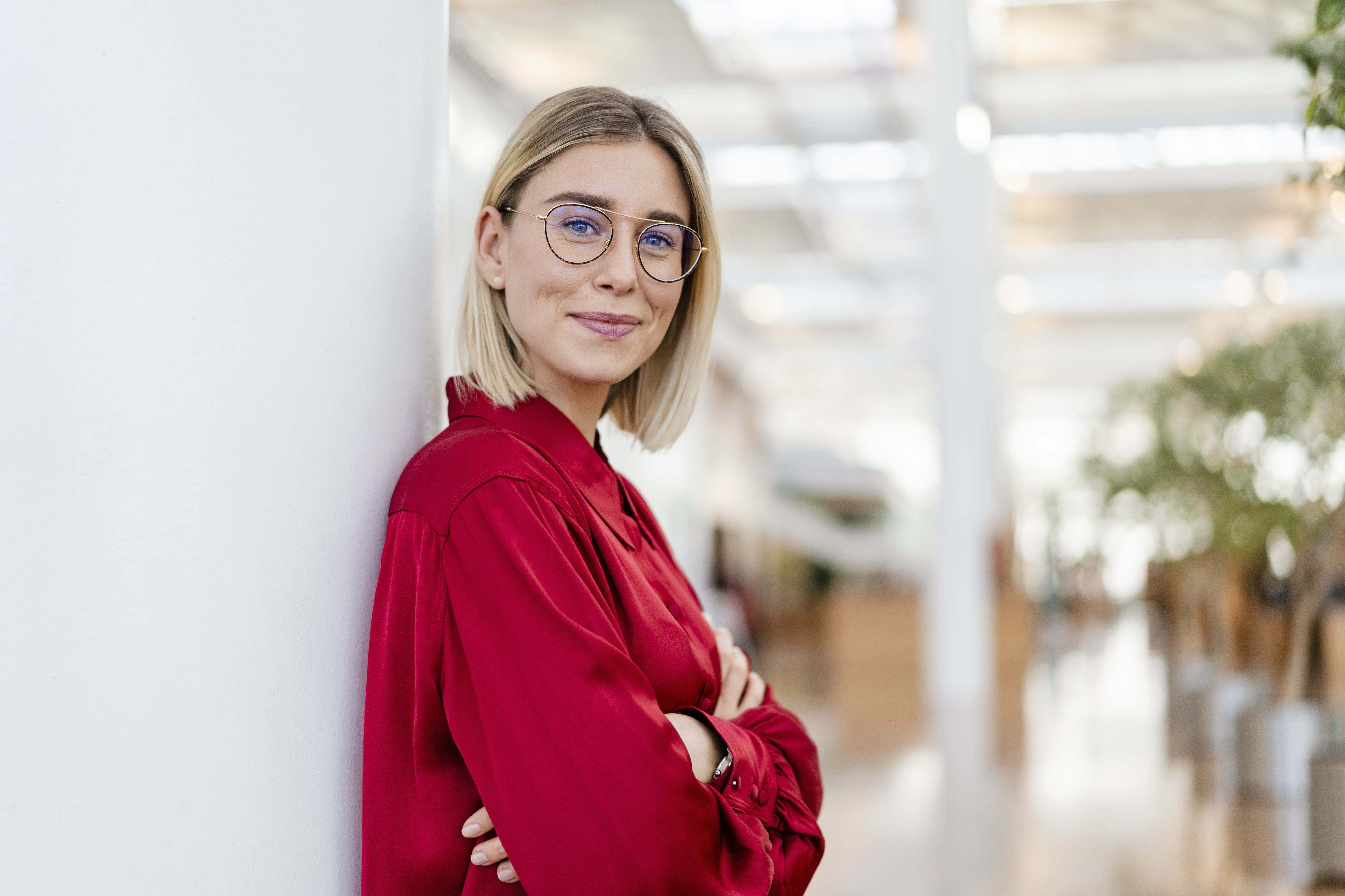 Eine junge Frau mit blonden, schulterlangen Haaren und Brille steht lächelnd an einer Wand. Sie trägt ein rotes Hemd und hat die Arme verschränkt. Der Hintergrund zeigt eine moderne, helle Innenarchitektur mit Pflanzen.