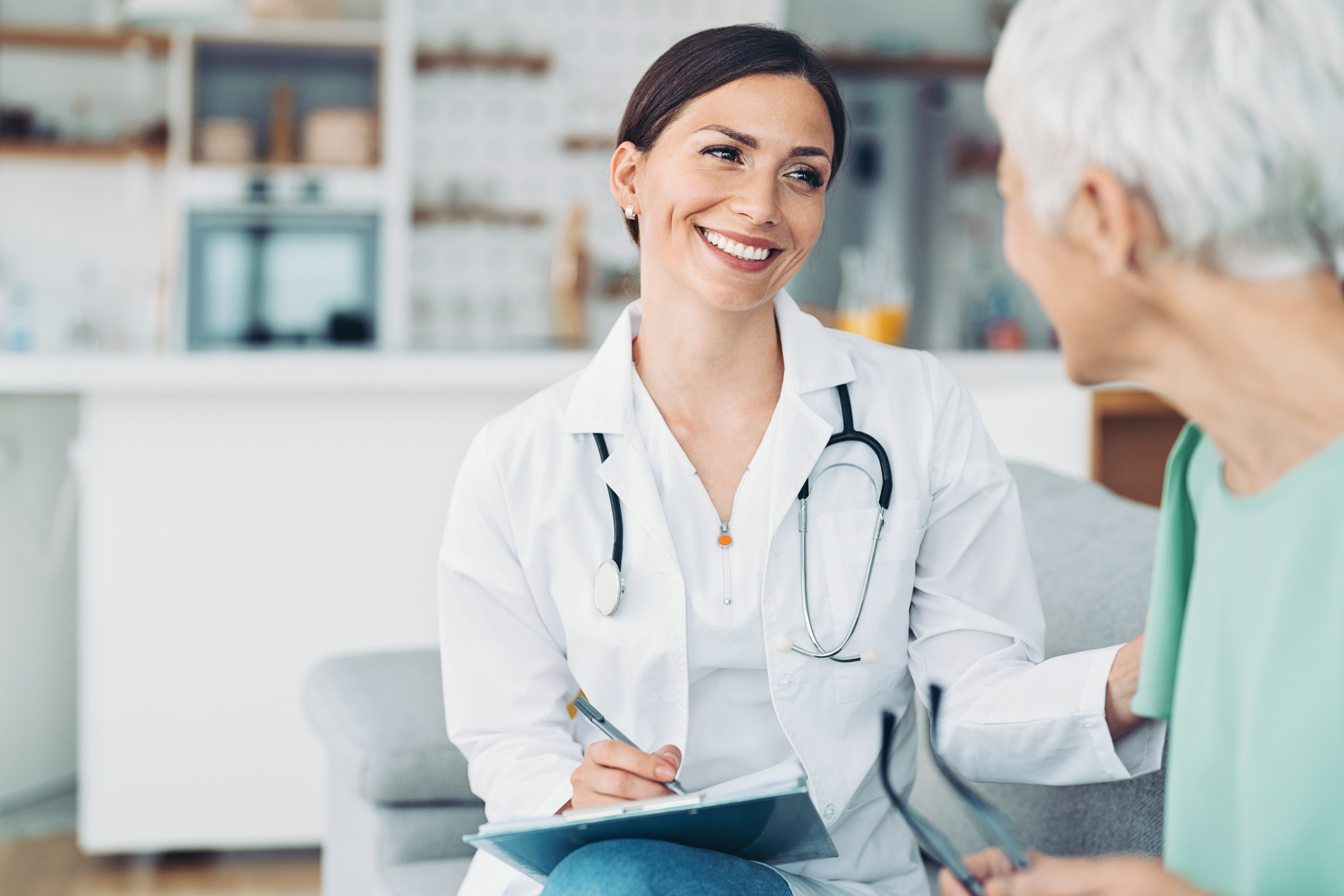 A doctor in a white coat is sitting on a sofa, talking to an elderly patient. The doctor is holding a notepad and smiling, while the patient listens attentively.