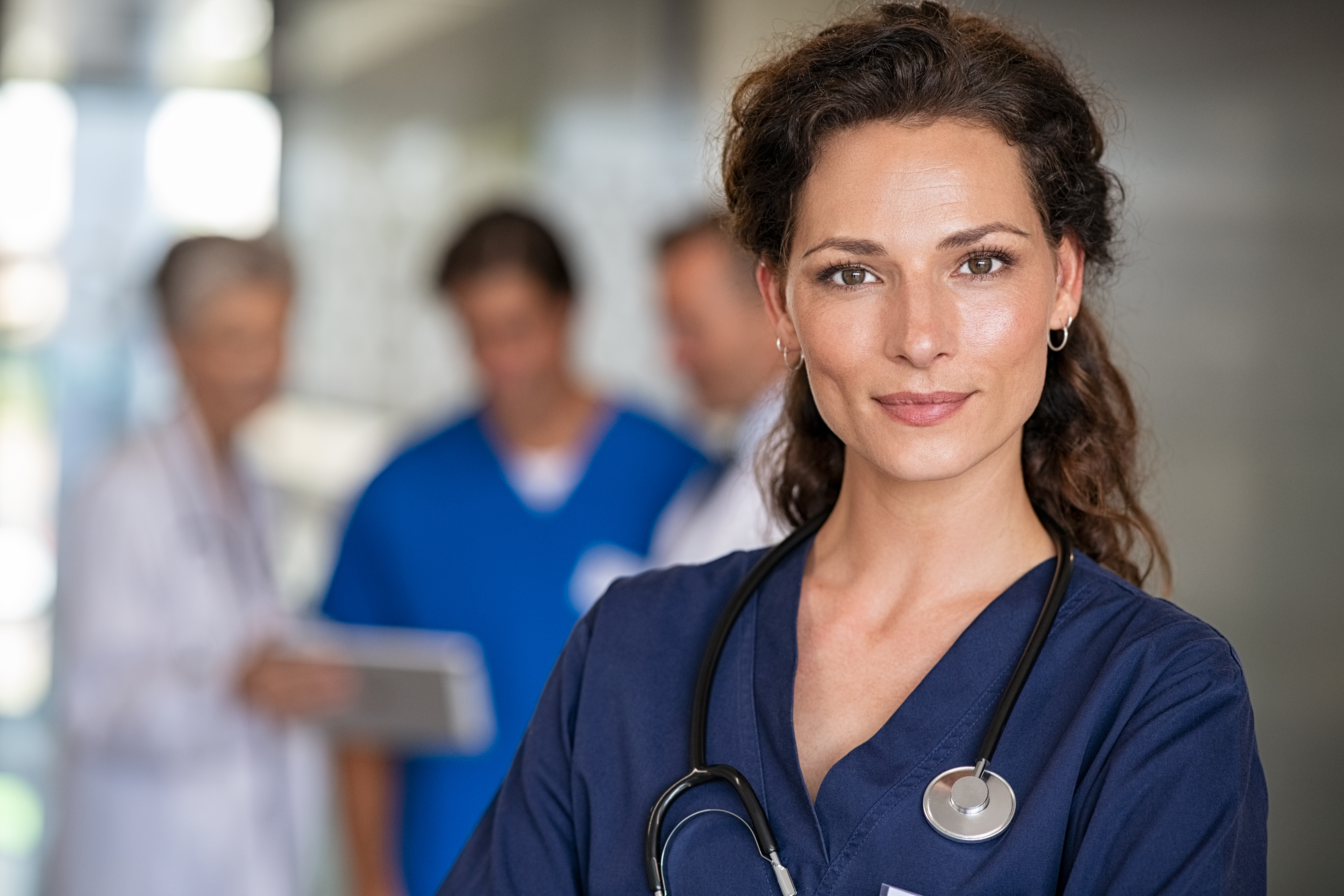 A female doctor in blue scrubs with a stethoscope around her neck stands in the foreground. In the background, two other medical professionals and a patient can be seen interacting with each other.