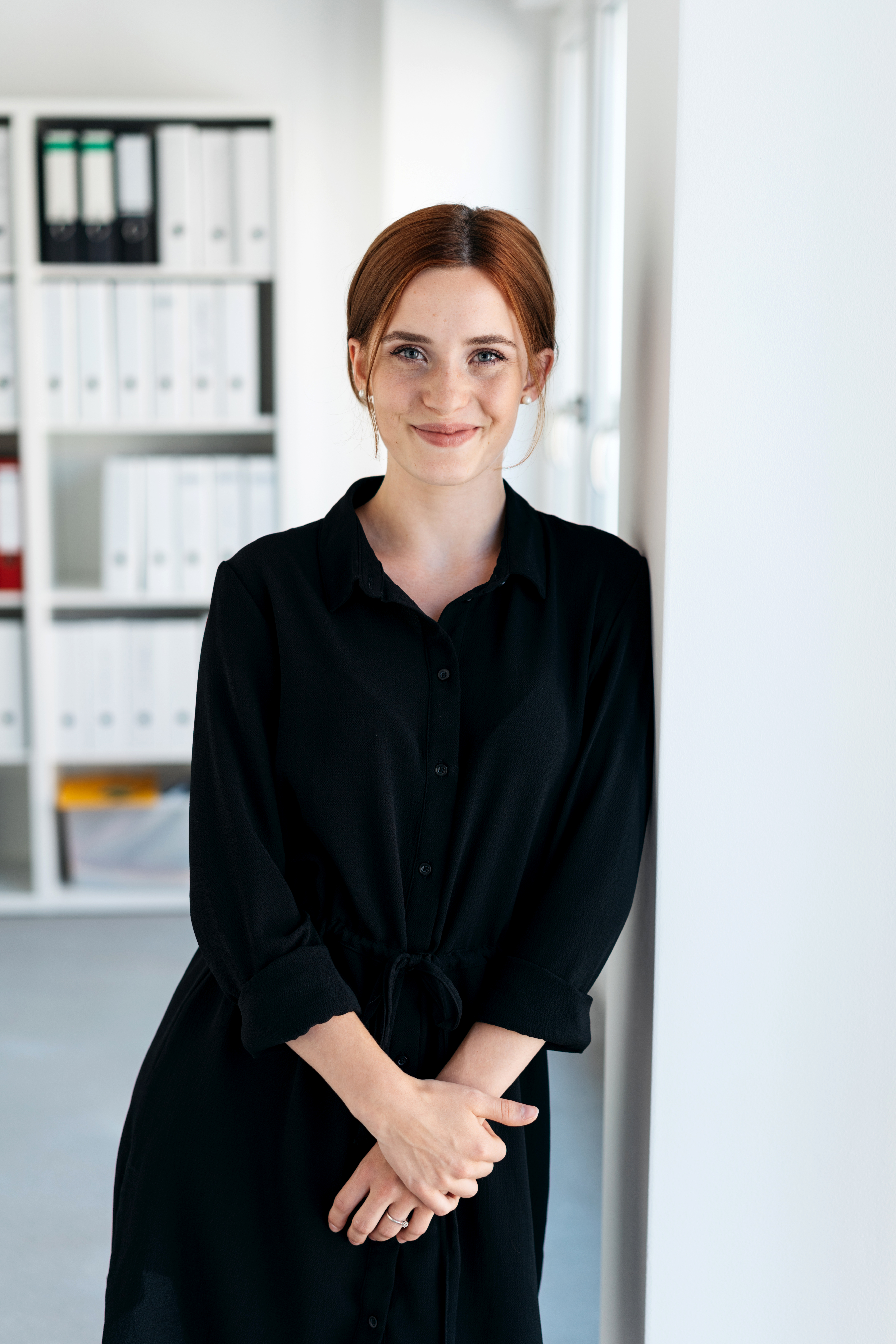 Eine Frau mit roten Haaren steht an einer Wand in einem modernen Büro. Sie trägt ein schwarzes Hemdkleid und lächelt. Im Hintergrund sind Regale mit Aktenordnern sichtbar.