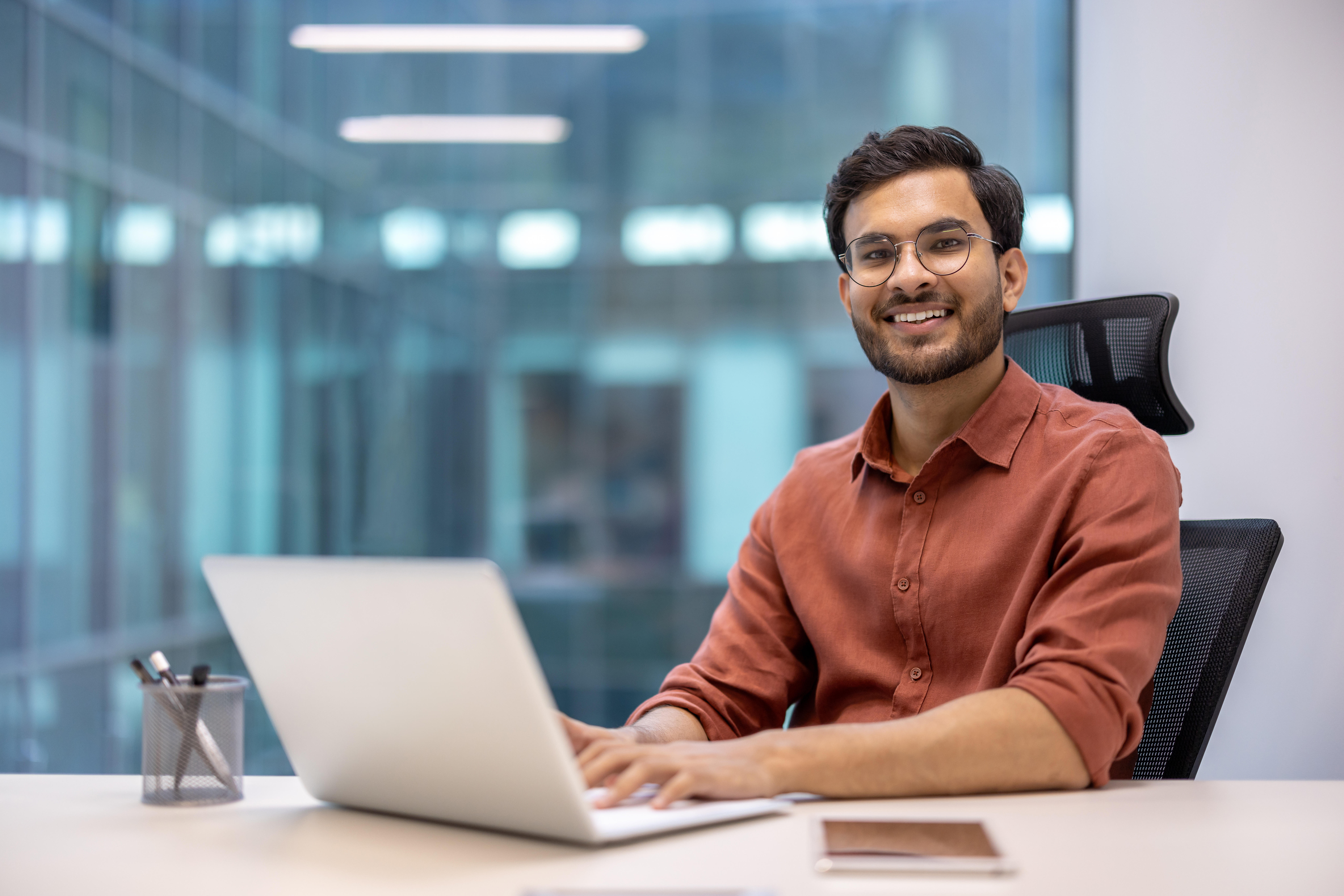 A man with glasses is sitting at a desk, working on a laptop. He is wearing a red shirt and smiling. On the desk, there is a pen holder and a notebook.