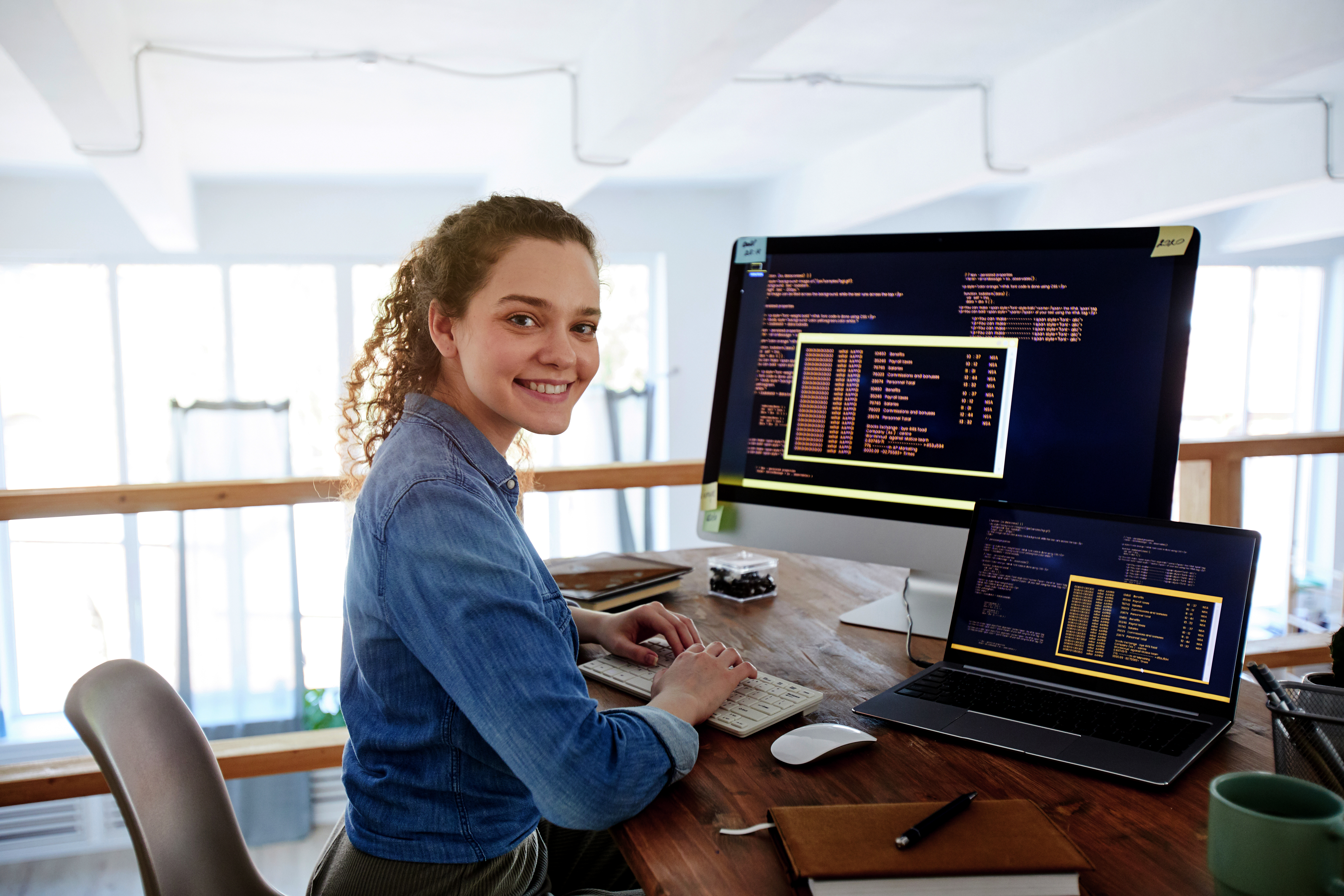 A young woman sits at a desk, working on two computers while smiling. Programming code is visible on the screens.