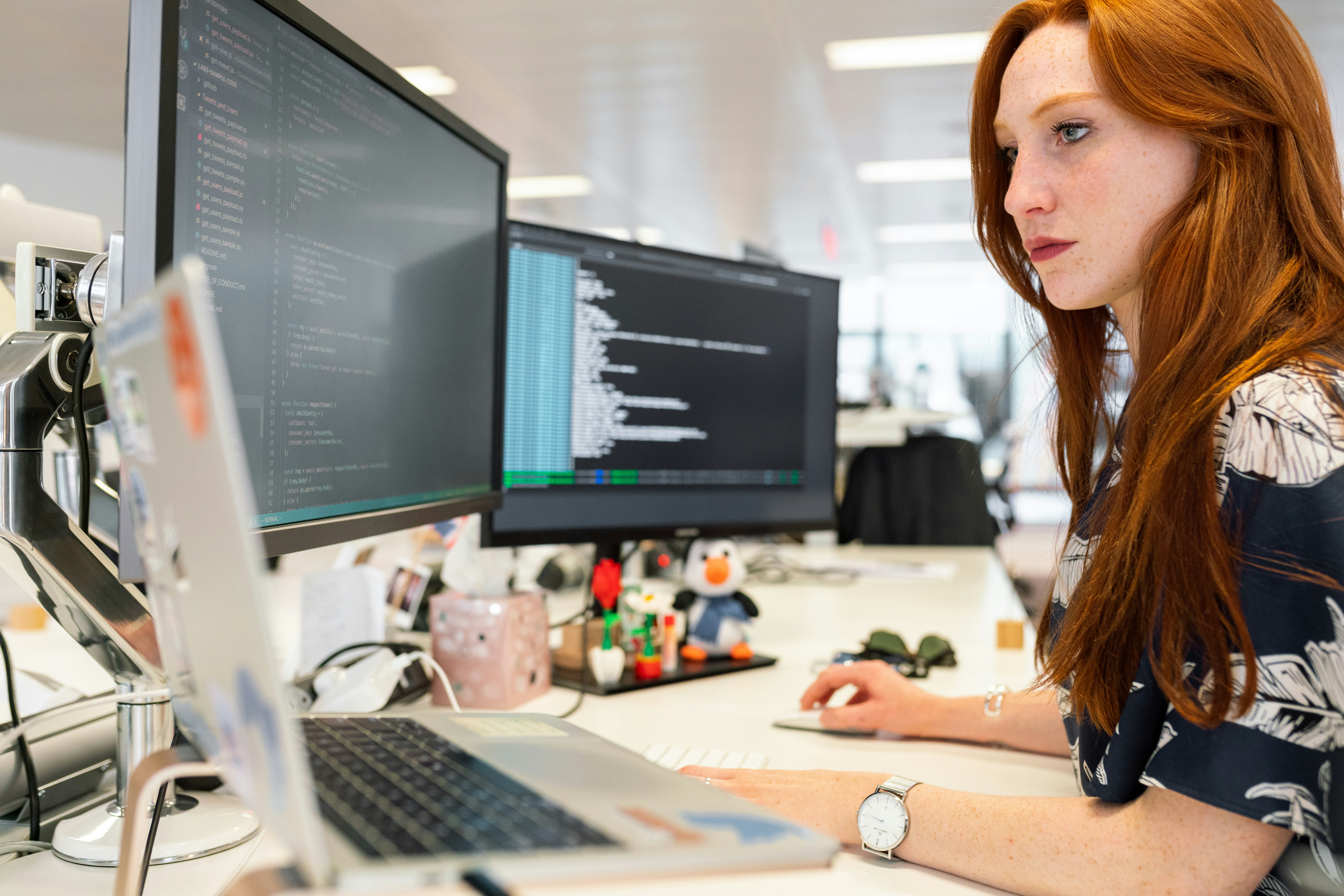 Eine Frau mit langen roten Haaren sitzt an einem Schreibtisch und arbeitet an einem Laptop. Vor ihr stehen zwei große Monitore mit Programmiercode. Der Arbeitsplatz ist modern und gut organisiert.