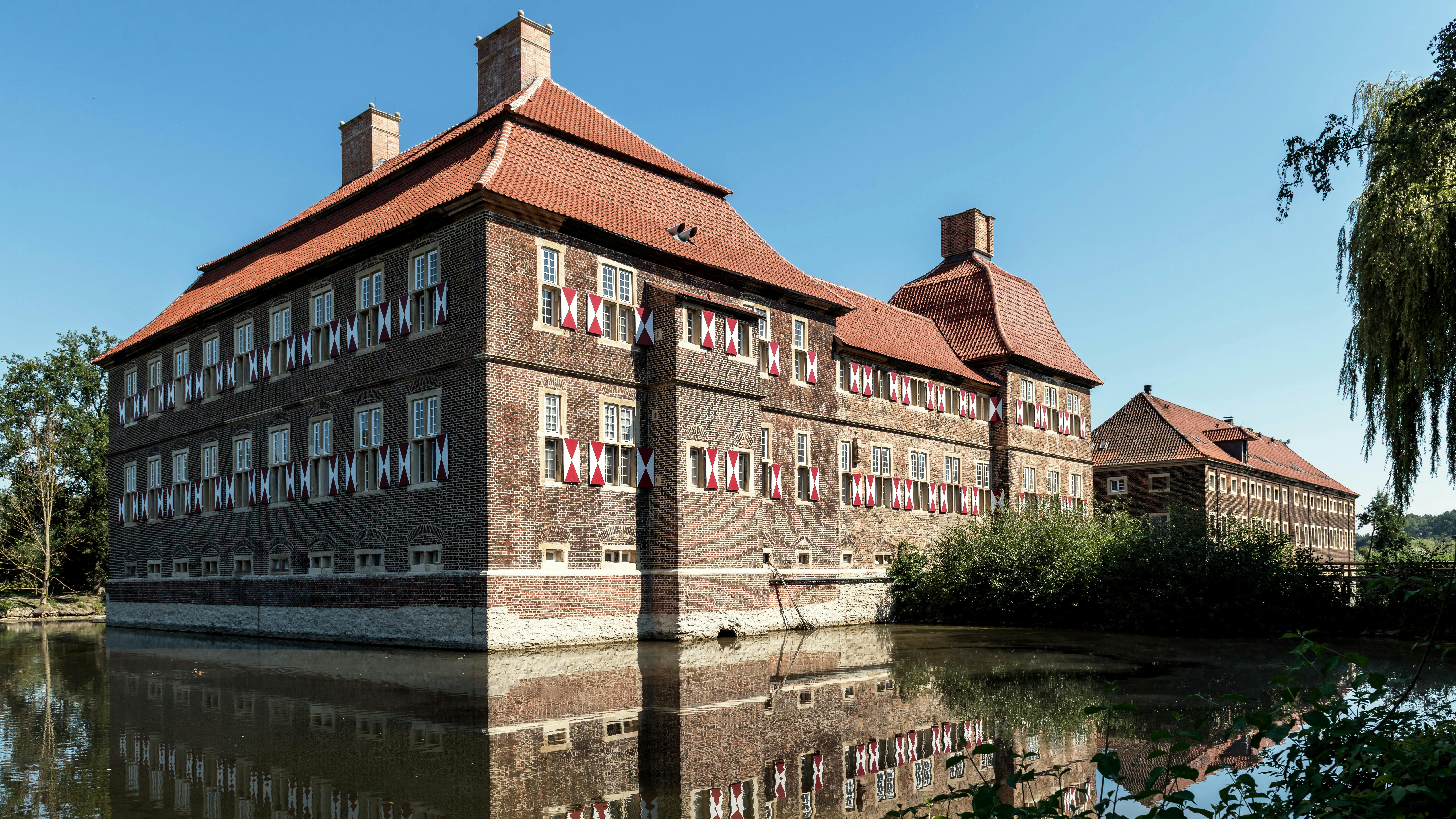 Historisches Gebäude mit rotem Satteldach und Fenstern mit bunten Vorhängen, umgeben von Wasser und Bäumen. Die Architektur zeigt eine Mischung aus Backstein und modernen Elementen.