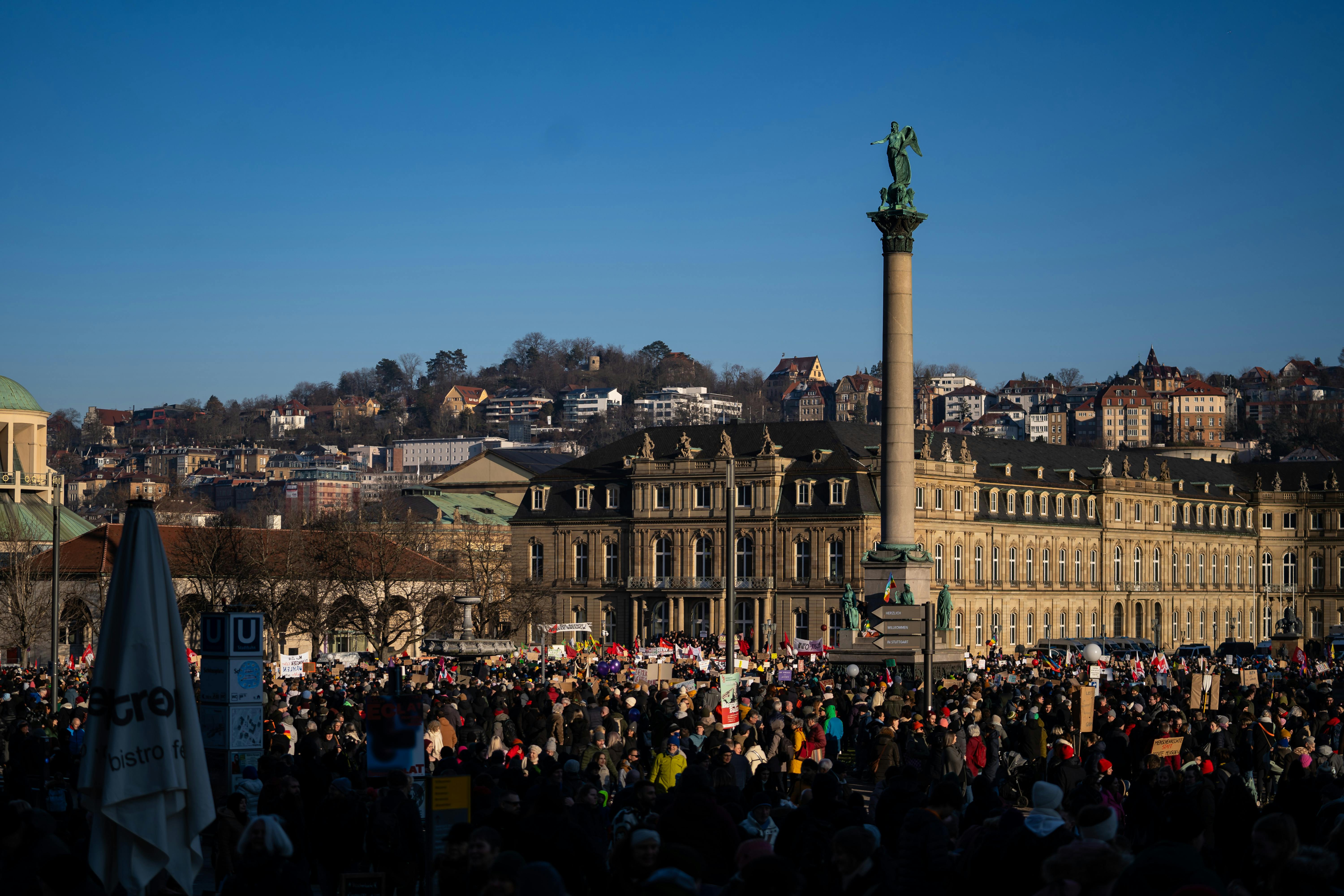 A large crowd gathers in a square, surrounded by historic buildings and a tall monument featuring a statue.