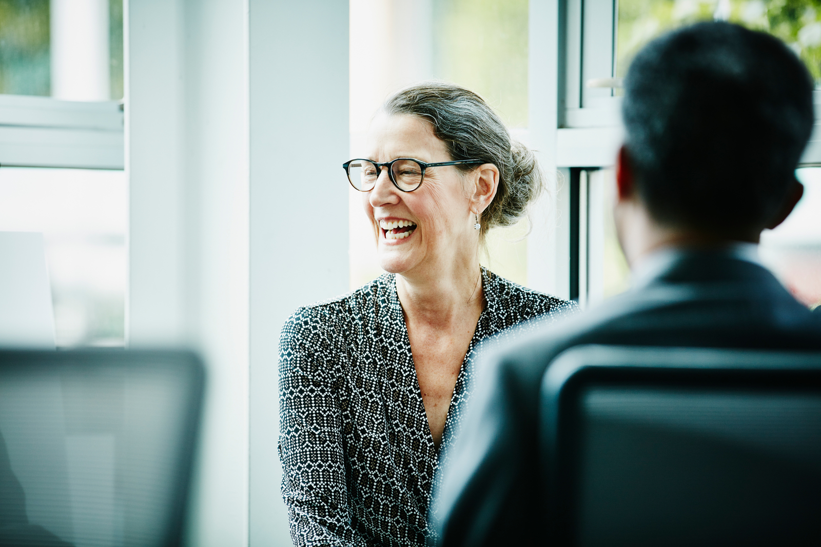 A smiling woman with glasses is wearing a patterned top and sitting in a modern office. In the background, another person can be seen sitting across from her.
