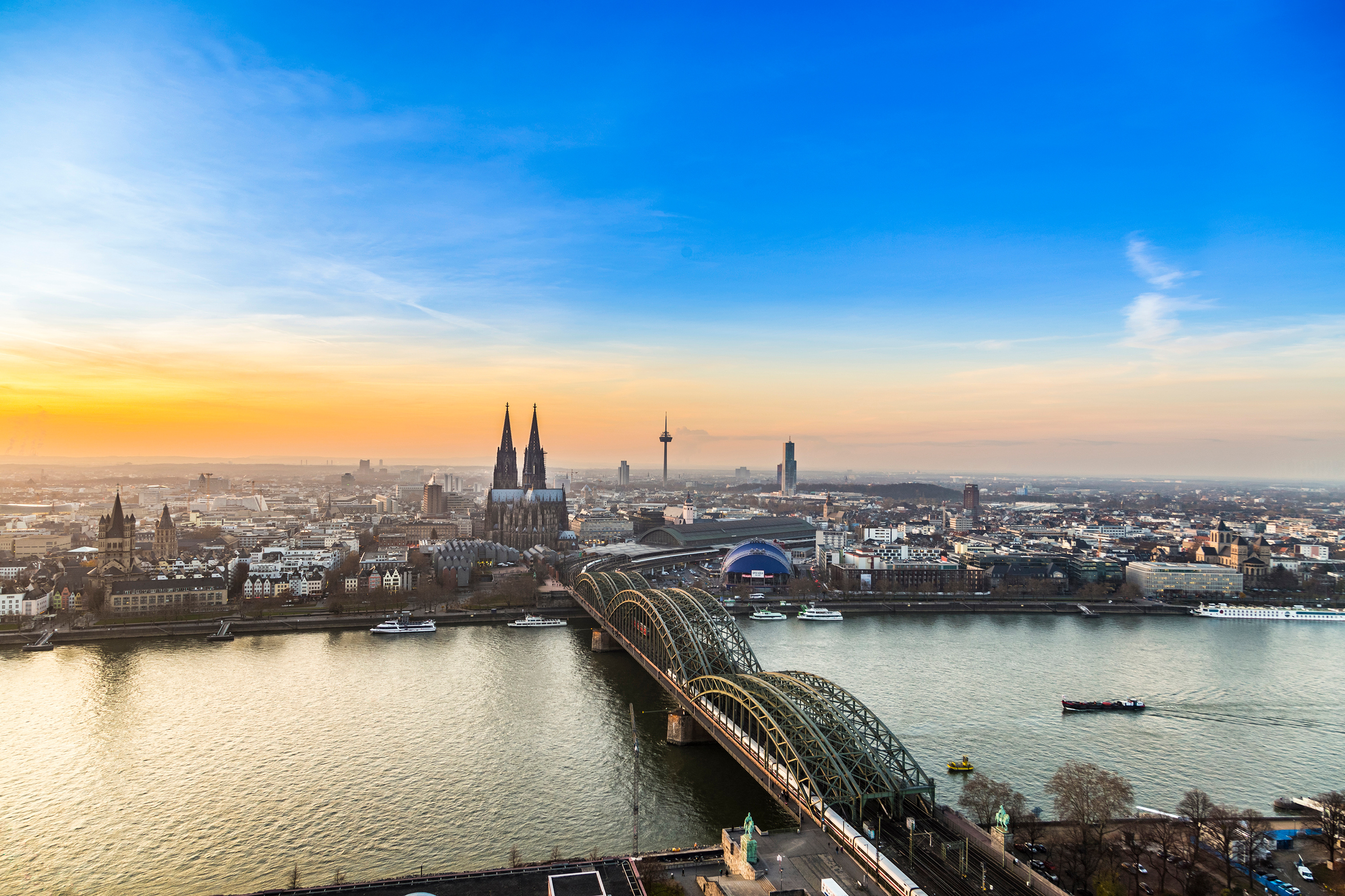Blick auf Köln bei Sonnenuntergang, mit der Hohenzollernbrücke und dem Kölner Dom im Vordergrund, während der Himmel in warmen Farben leuchtet.