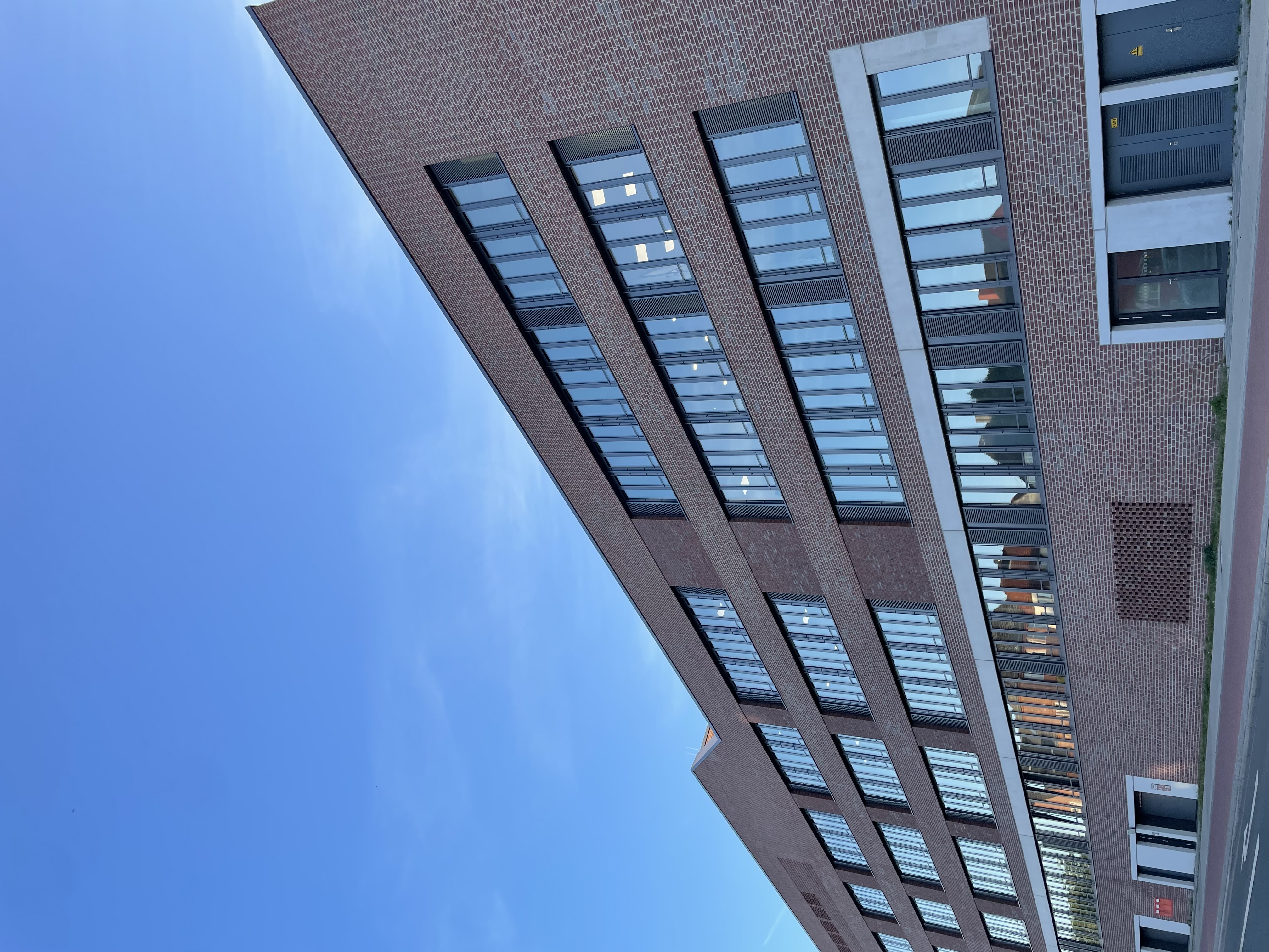 Moderne Backsteinfassade eines mehrstöckigen Gebäudes mit großen Fenstern und klaren Linien, unter einem blauen Himmel.
