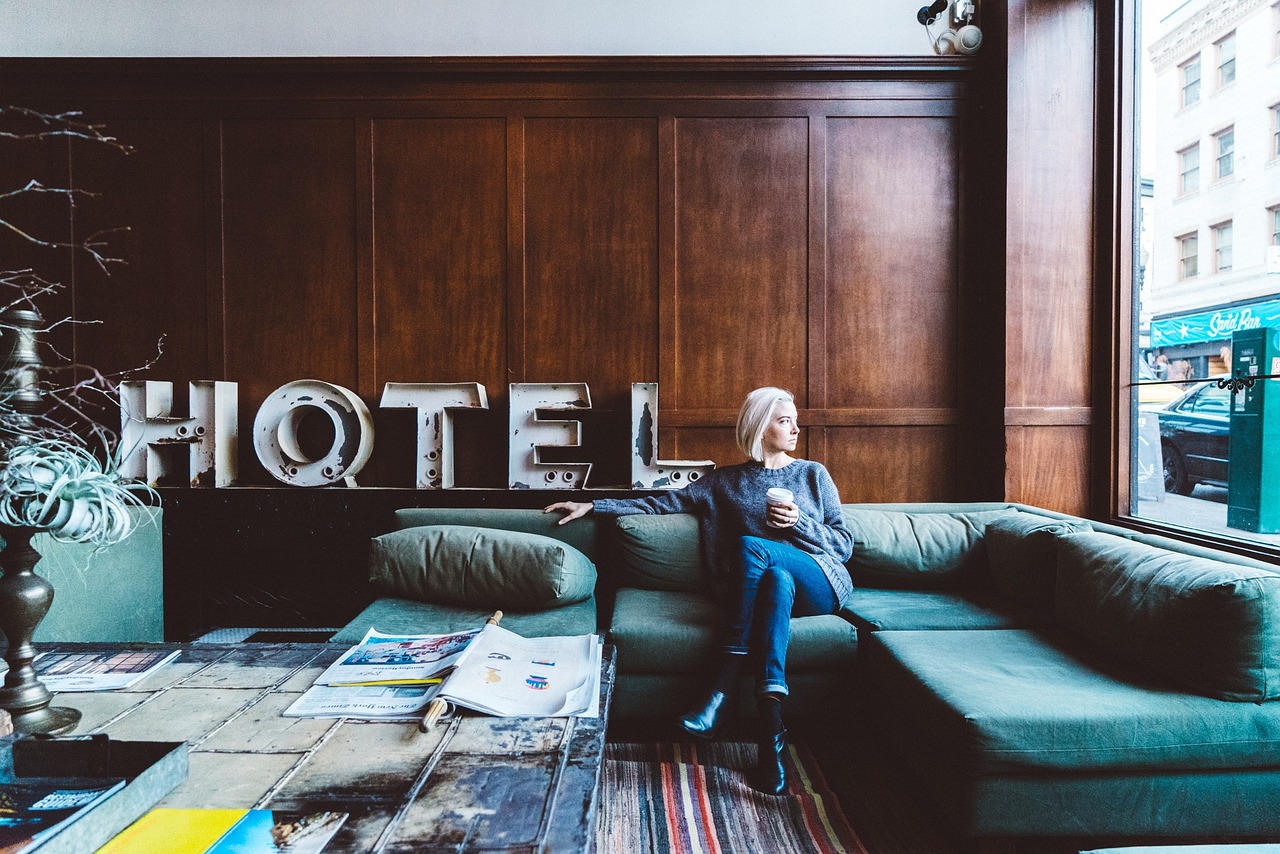 A woman with blonde hair is sitting on a green sofa in a hotel lobby. She holds a cup in her hand and gazes thoughtfully out the window. In the background, the word "HOTEL" is displayed in large letters.
