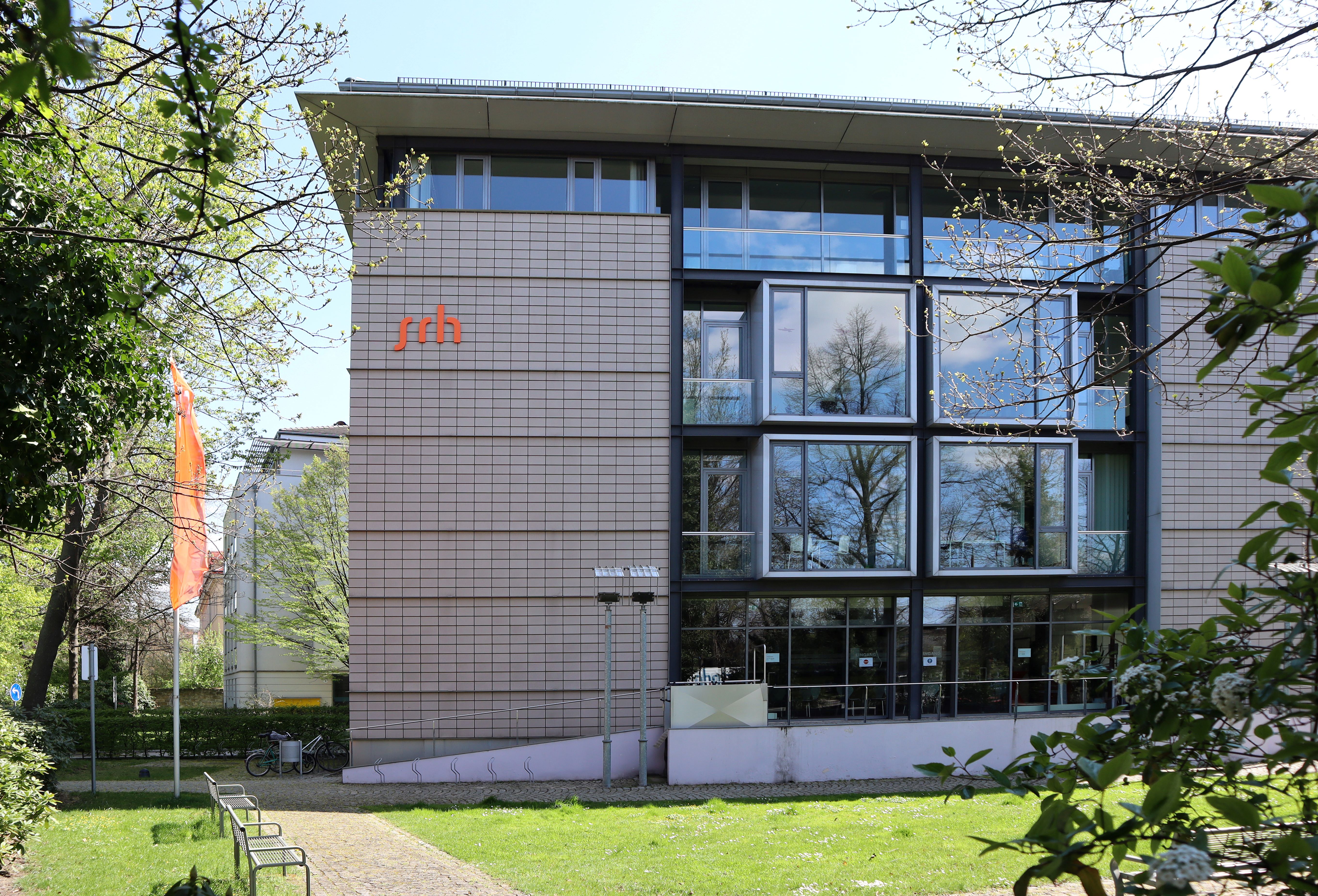 Moderne Gebäude mit großen Fenstern, beschriftet mit "srh", umgeben von Bäumen und Grünflächen.