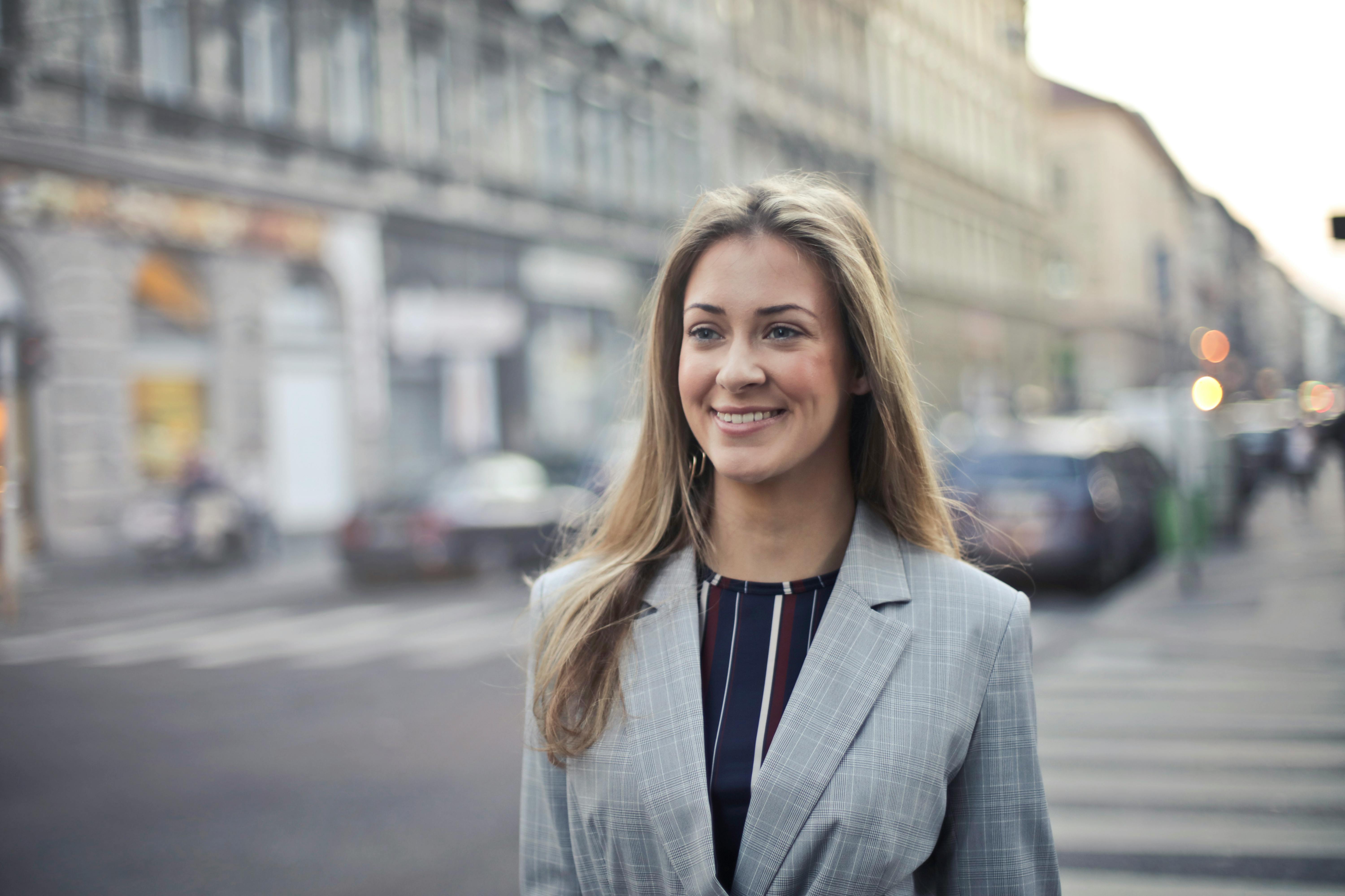 A smiling woman with long, curly hair is wearing a gray suit and standing on a street, surrounded by buildings and cars in the background.