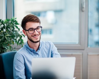 Ein junger Mann mit Brille sitzt an einem Schreibtisch und lächelt, während er auf einen Laptop schaut. Im Hintergrund ist eine Pflanze und ein Fenster sichtbar.