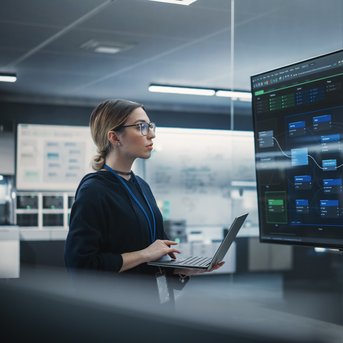 A woman stands in front of a large screen displaying data and graphics, while holding a laptop in her hand.