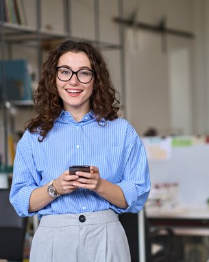 A woman with curly hair and glasses stands in a modern office, holding a smartphone in her hand. She is wearing a striped shirt.