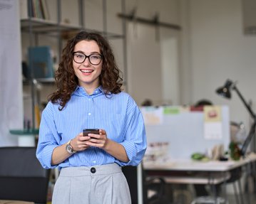 Eine Frau mit lockigem Haar und Brille steht in einem modernen Büro und hält ein Smartphone in der Hand. Sie trägt ein gestreiftes Hemd.