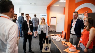 Eine Gruppe von fünf Personen steht an einem Tisch mit Präsentationsmaterialien und Snacks, während sie miteinander sprechen.