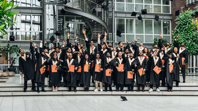 A group of graduates in black gowns, throwing their caps in the air and holding their diplomas in their hands, stands in a modern building.