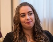 Eine Frau mit lockigem, braunem Haar sitzt in einem Raum und schaut aufmerksam. Sie trägt eine schwarze Jacke und hat einen neutralen Gesichtsausdruck.