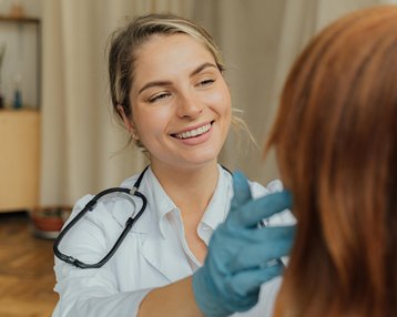 Eine lächelnde Ärztin untersucht das Haar einer Patientin. Die Ärztin trägt Handschuhe und hat ein Stethoskop um den Hals. Im Hintergrund sind einige Möbel und Pflanzen sichtbar.