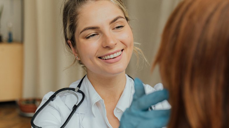 Eine lächelnde Ärztin untersucht das Haar einer Patientin. Die Ärztin trägt Handschuhe und hat ein Stethoskop um den Hals. Im Hintergrund sind einige Möbel und Pflanzen sichtbar.