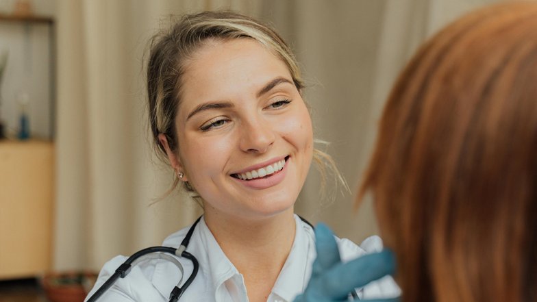 Eine lächelnde Ärztin untersucht das Haar einer Patientin. Die Ärztin trägt Handschuhe und hat ein Stethoskop um den Hals. Im Hintergrund sind einige Möbel und Pflanzen sichtbar.