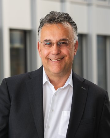 Ein Mann in einem schwarzen Anzug mit einem weißen Hemd lächelt freundlich. Er steht vor einem modernen Hintergrund mit Fenstern.