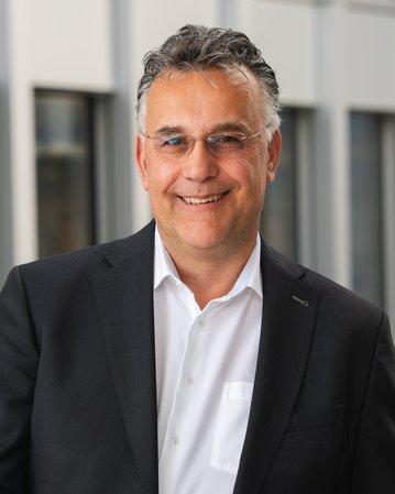 A man in a black suit with a white shirt smiles warmly. He stands in front of a modern backdrop with windows.