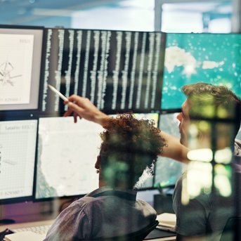 Zwei Personen betrachten mehrere Computerbildschirme mit technischen Daten und Grafiken, während sie gemeinsam an einem Projekt arbeiten.