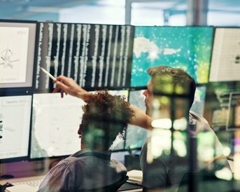 Zwei Personen betrachten mehrere Computerbildschirme mit technischen Daten und Grafiken, während sie gemeinsam an einem Projekt arbeiten.