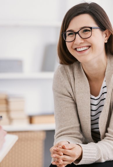 Eine lächelnde Frau mit Brille sitzt in einem Gespräch, während eine andere Person, die nicht sichtbar ist, Notizen macht. Die Umgebung wirkt freundlich und modern.